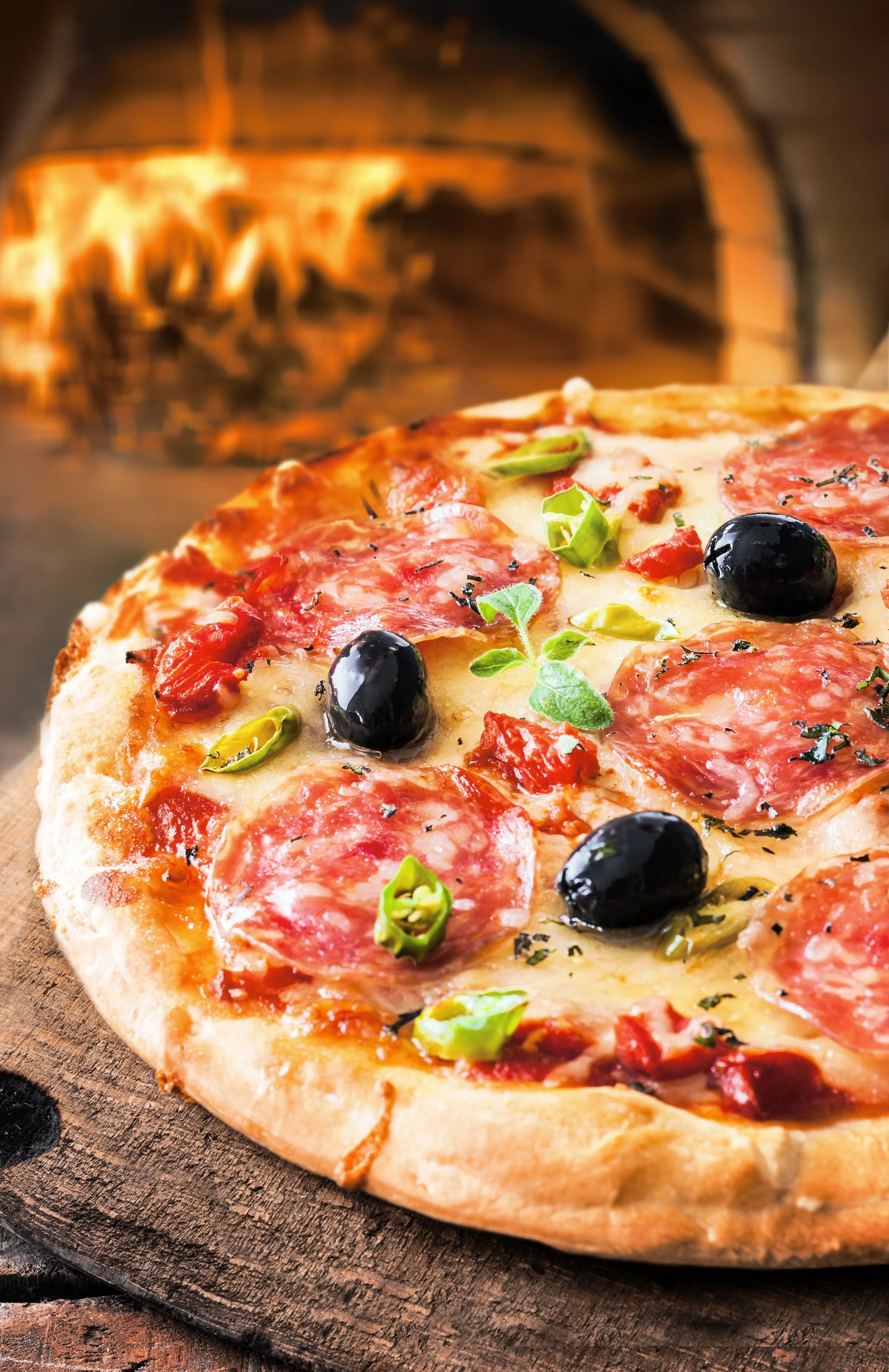 Close-up of a pizza with pepperoni, green peppers, black olives, herbs, and melted cheese in front of a brick oven.