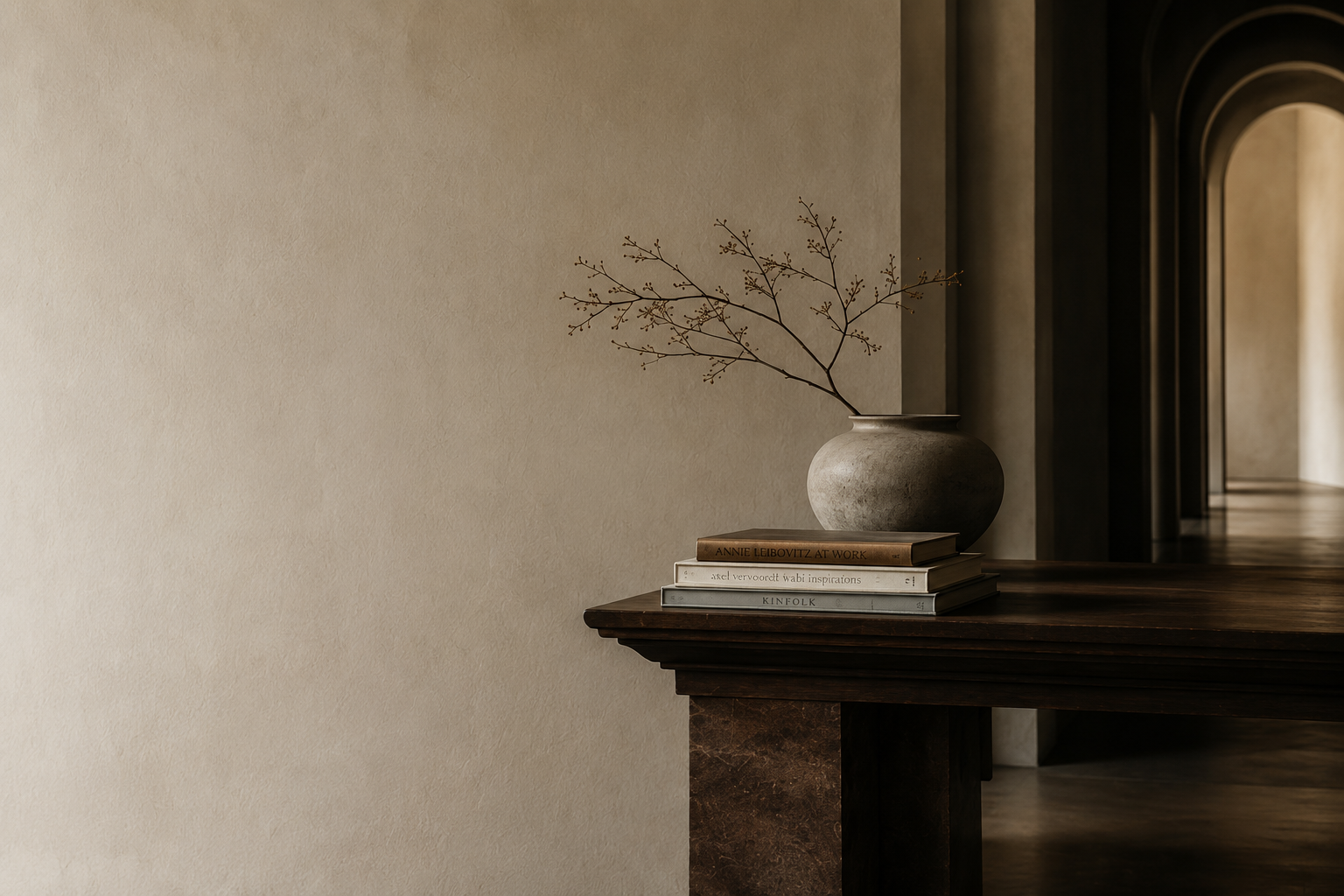 A minimalist interior with a neutral-colored wall and a wooden console table. On the table, there are stacked books and a round, textured vase containing a thin, leafless branch.
