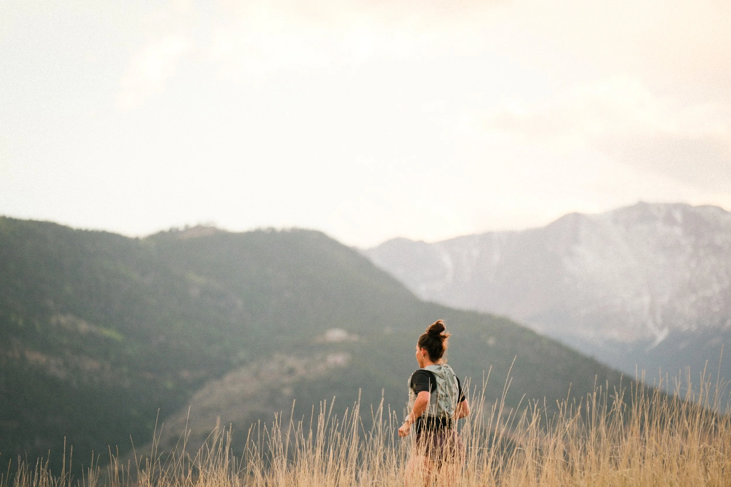 A woman with a backpack standing in a field of tall grass, looking towards distant mountains under a cloudy sky.