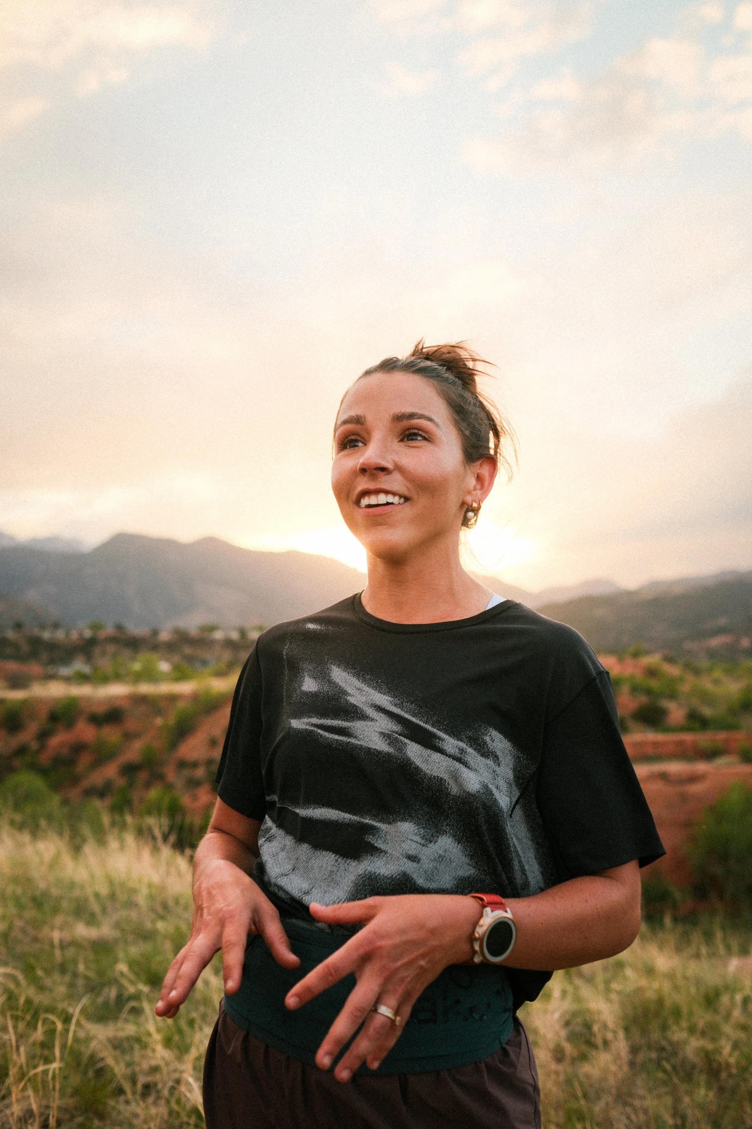 Woman smiling outdoors at sunset, standing in a grassy field with mountains in the background, wearing a black T-shirt, smartwatch, earrings, and rings.