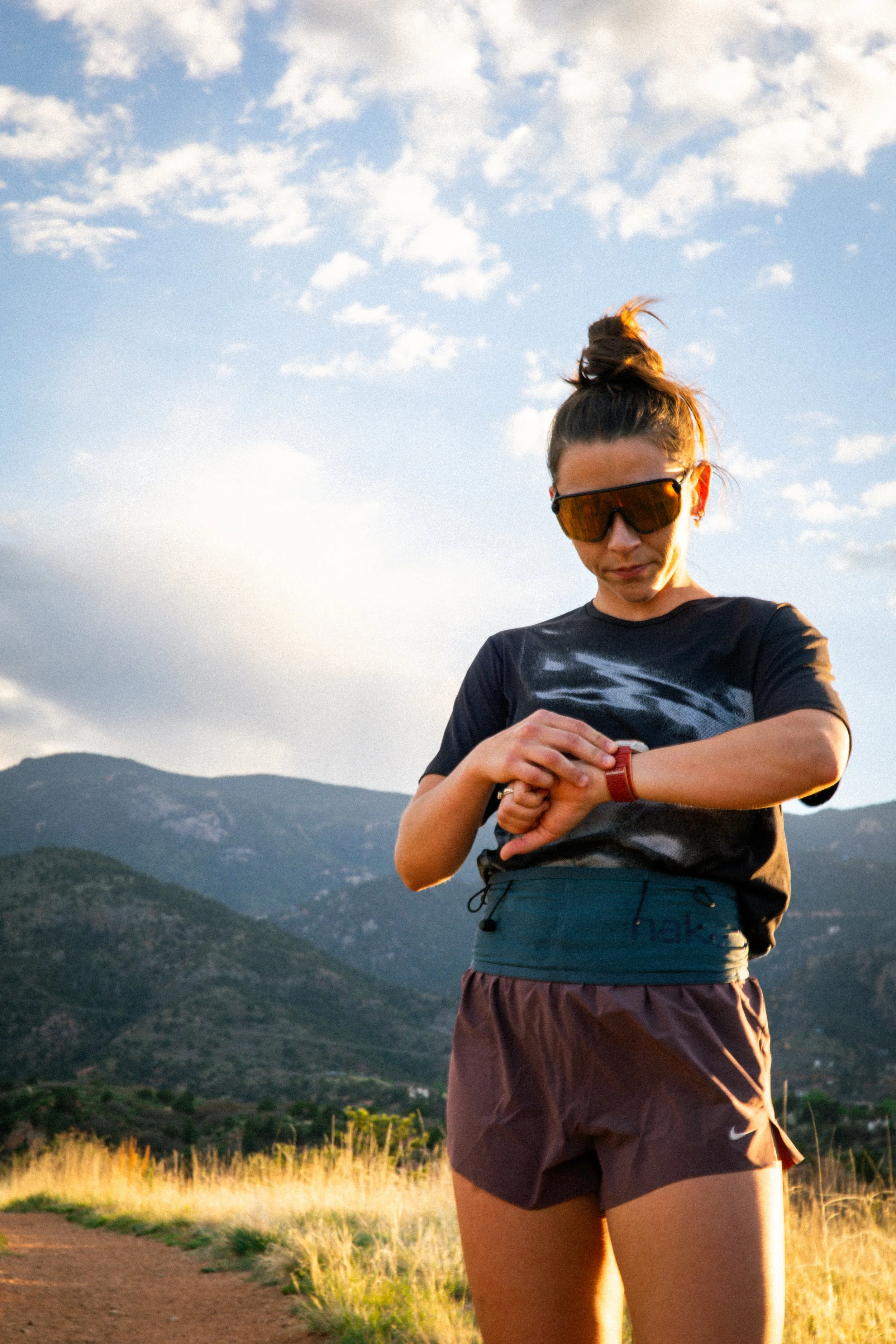 A woman wearing sunglasses and athletic clothing standing outdoors in a mountainous landscape during sunset, checking her watch.