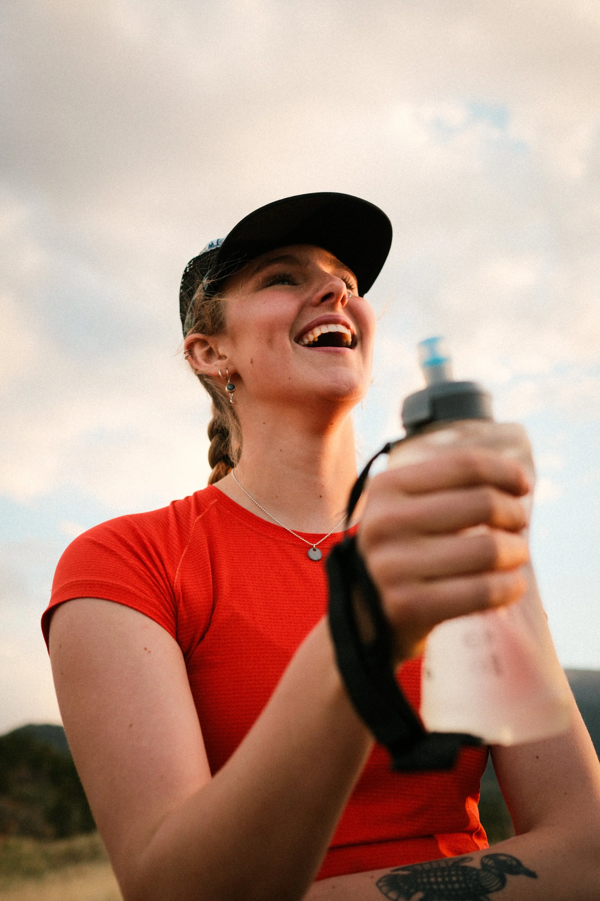 A young woman with a braid, earrings, and a necklace, smiling and holding a water bottle outdoors in the evening.