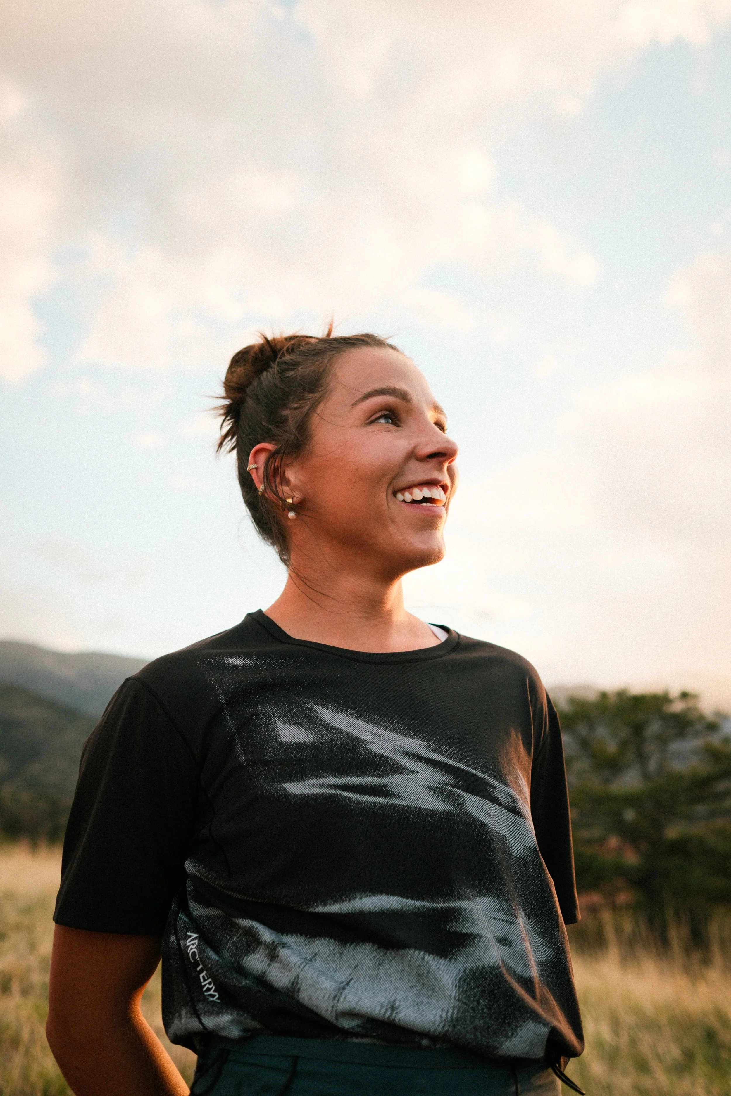 A young woman with short brown hair in a bun, wearing pearl earrings and a black athletic shirt, smiling and looking to the side outdoors during sunset with a cloudy sky and mountains in the background.