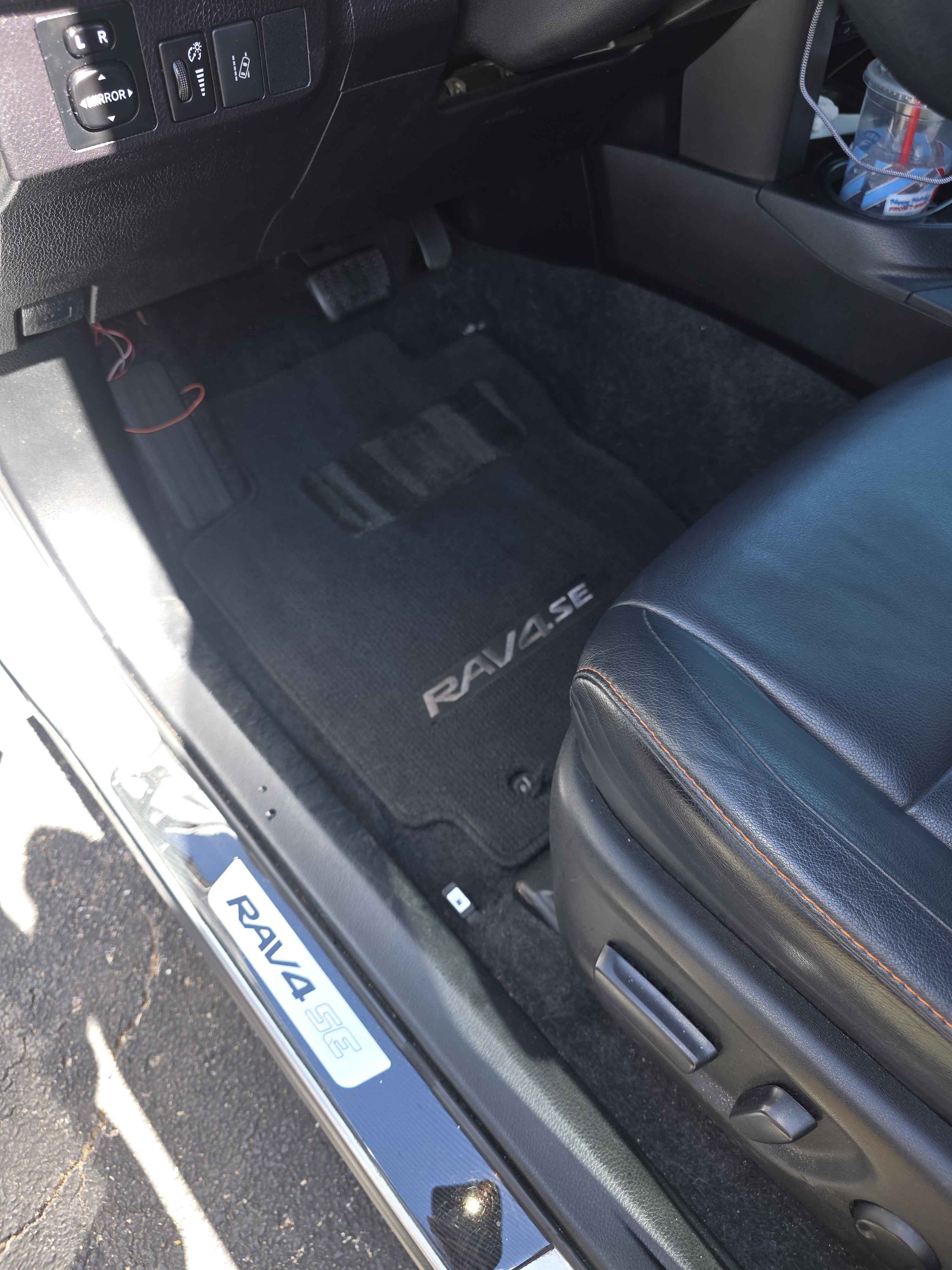 Inside a Toyota RAV4 SE, showing the driver's side footwell with a black floor mat, pedals, and part of the seat with orange stitching. The door sill has a silver plate with the RAV4 SE logo, and the cup holder on the right contains a plastic cup with a straw.