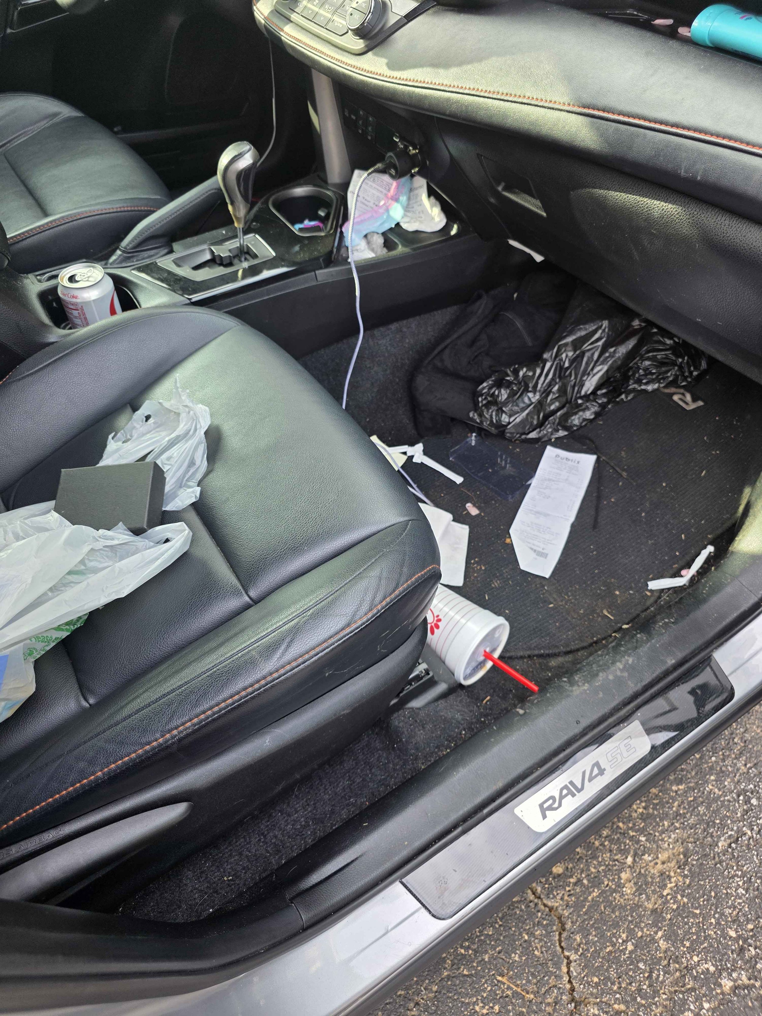 The interior of a car with clutter including a tear-out receipt, trash, a drink cup, used tissues, and a Coca-Cola can, with a gear shift and dashboard visible.