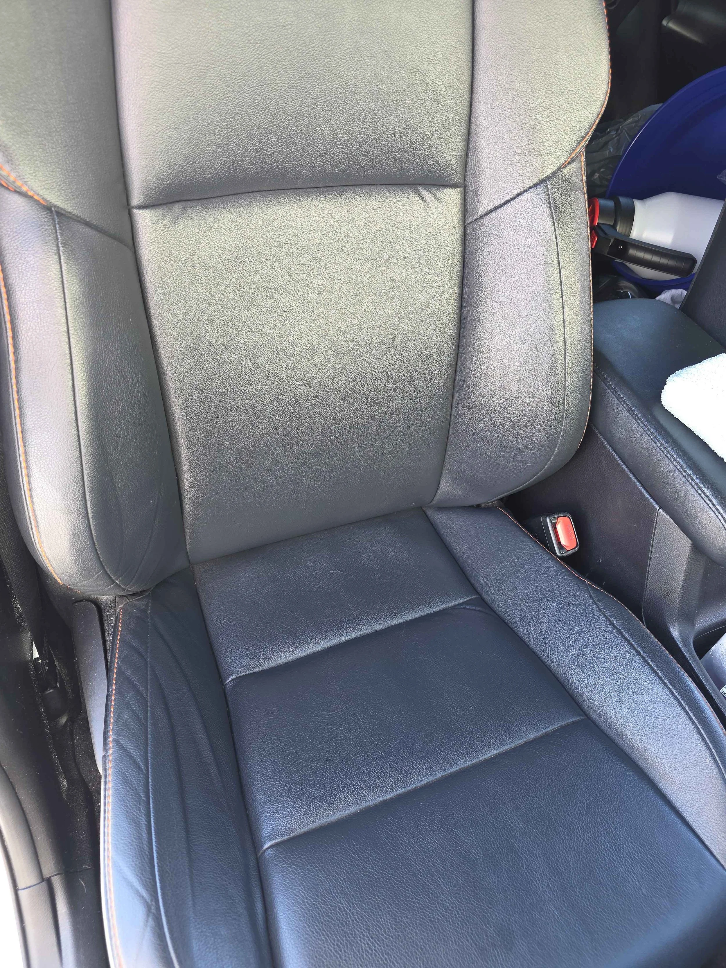 A black leather car seat with orange contrast stitching, showing the backrest and seat cushion, with a seatbelt buckle visible to the right.