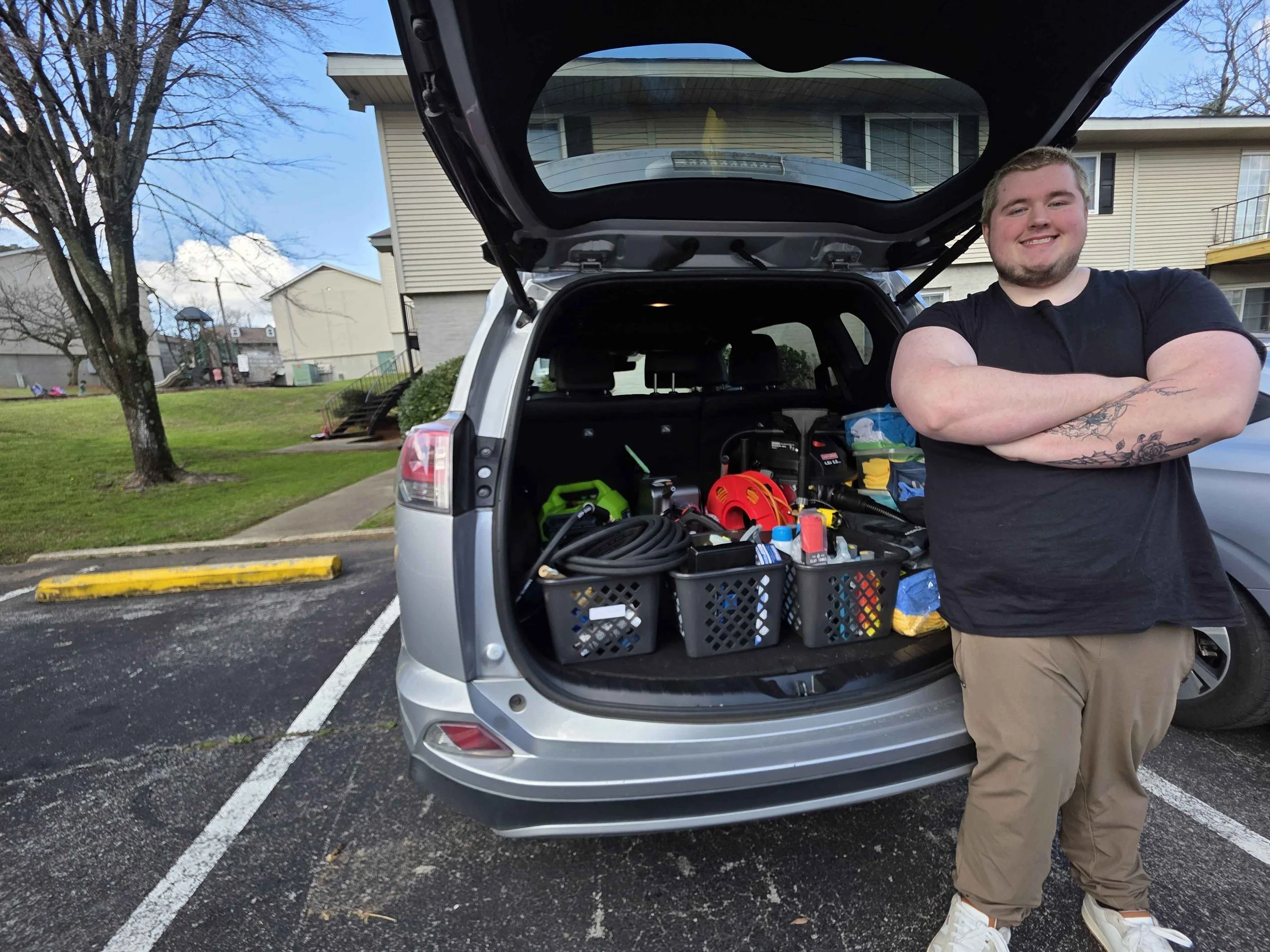 A young man with crossed arms standing next to an open car trunk filled with tools and supplies in a residential parking lot.