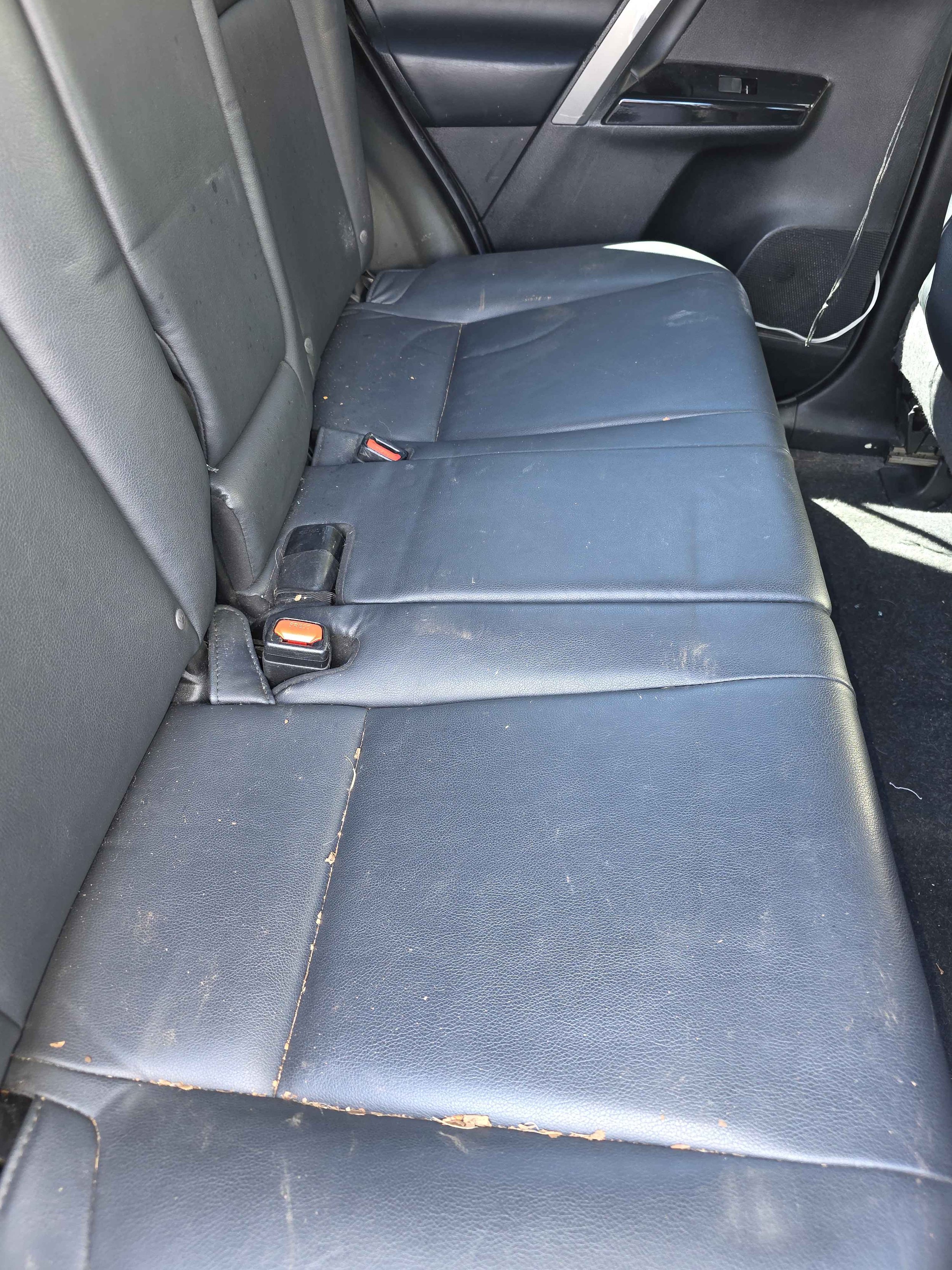 Back seat of a car with black leather upholstery, showing signs of wear, scratches, and dirt, with two visible seatbelt buckles.