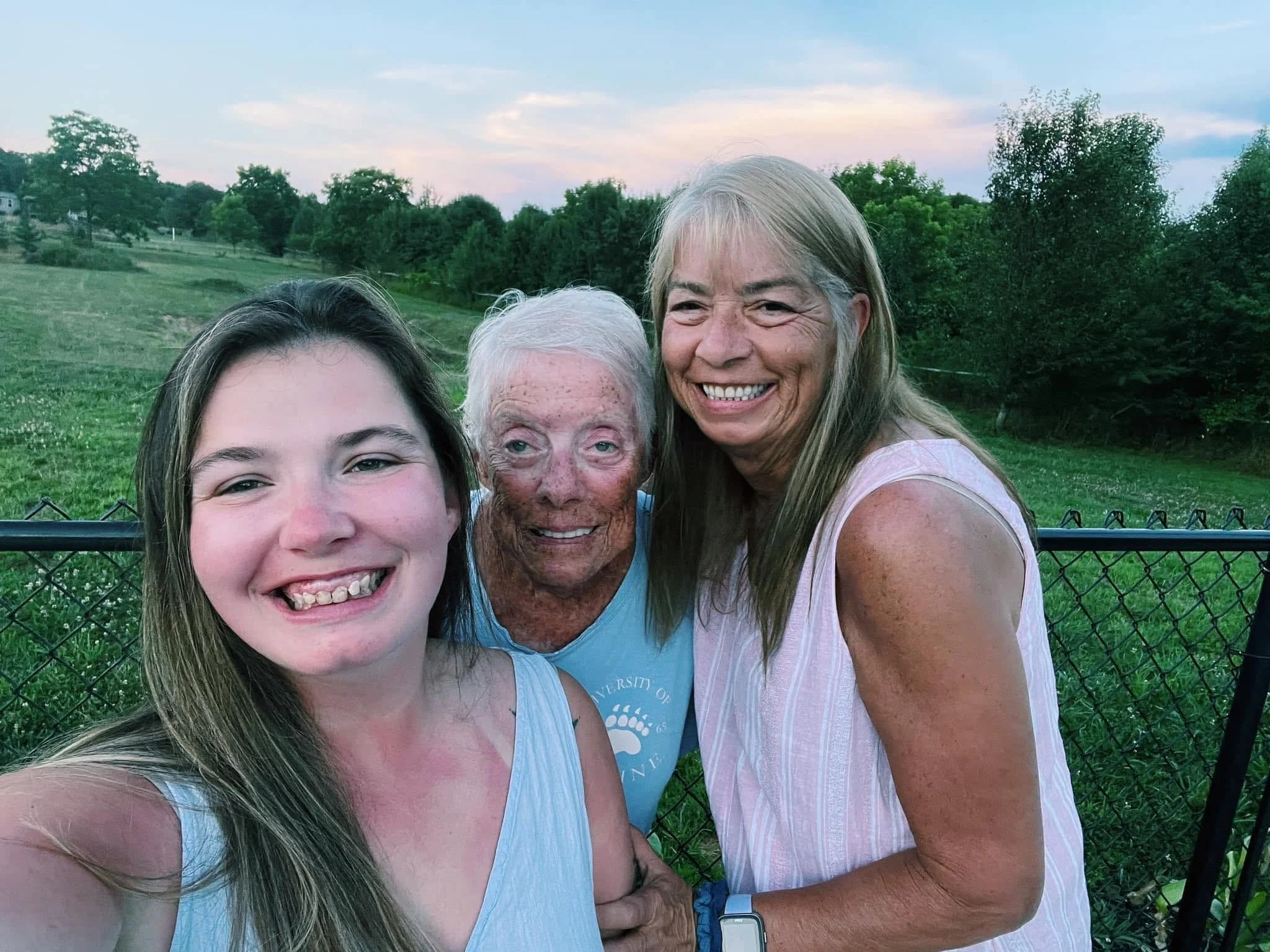 Four women smiling outdoors, two older and two younger, in a green field with trees and a fence in the background during sunset.