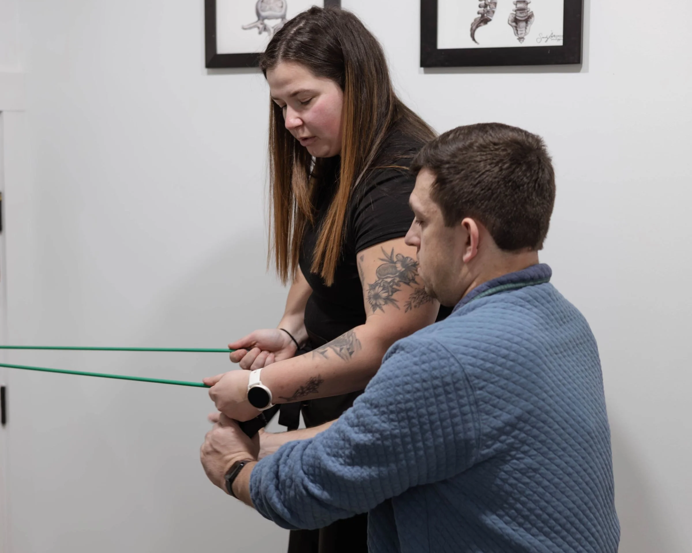 A woman with long brown hair and tattoos on her arm is assisting a man with short dark hair, wearing a blue quilted jacket, as they both hold green poles in a room with white walls and framed artwork of insects hanging behind them.