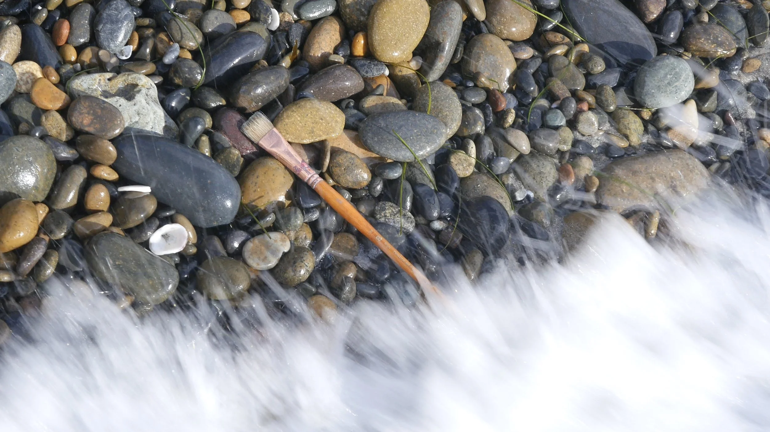 A close-up of smooth, wet rocks of various sizes and colors at water's edge along a shoreline, with a paintbrush among the rocks.