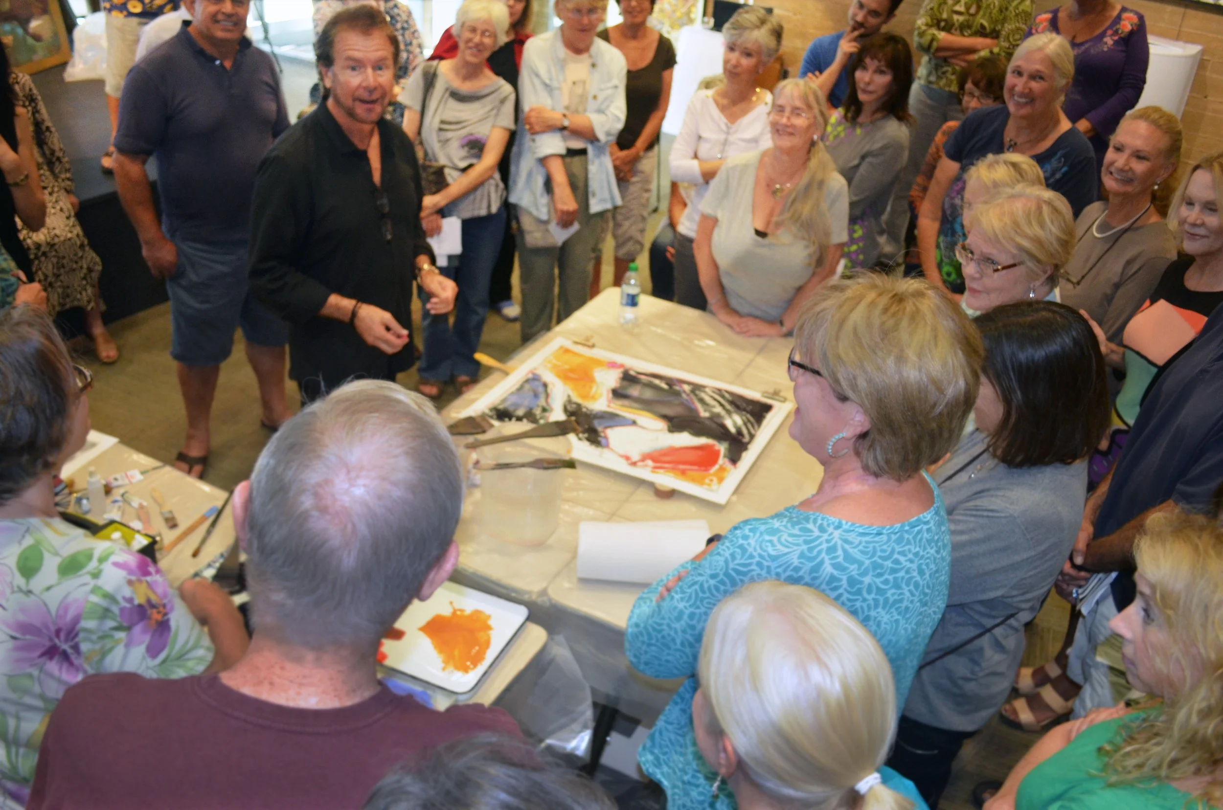 A man explaining and demonstrating to a group of people gathered around a table with an abstract painting and art supplies in a lively indoor setting.