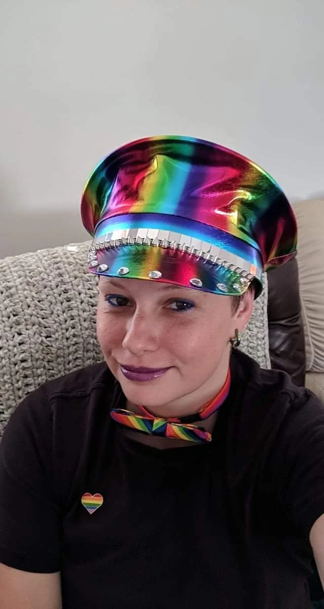 Person wearing a rainbow-colored hat, a matching rainbow bowtie, and a black shirt with a rainbow heart pin, sitting on a sofa with a beige cushion.