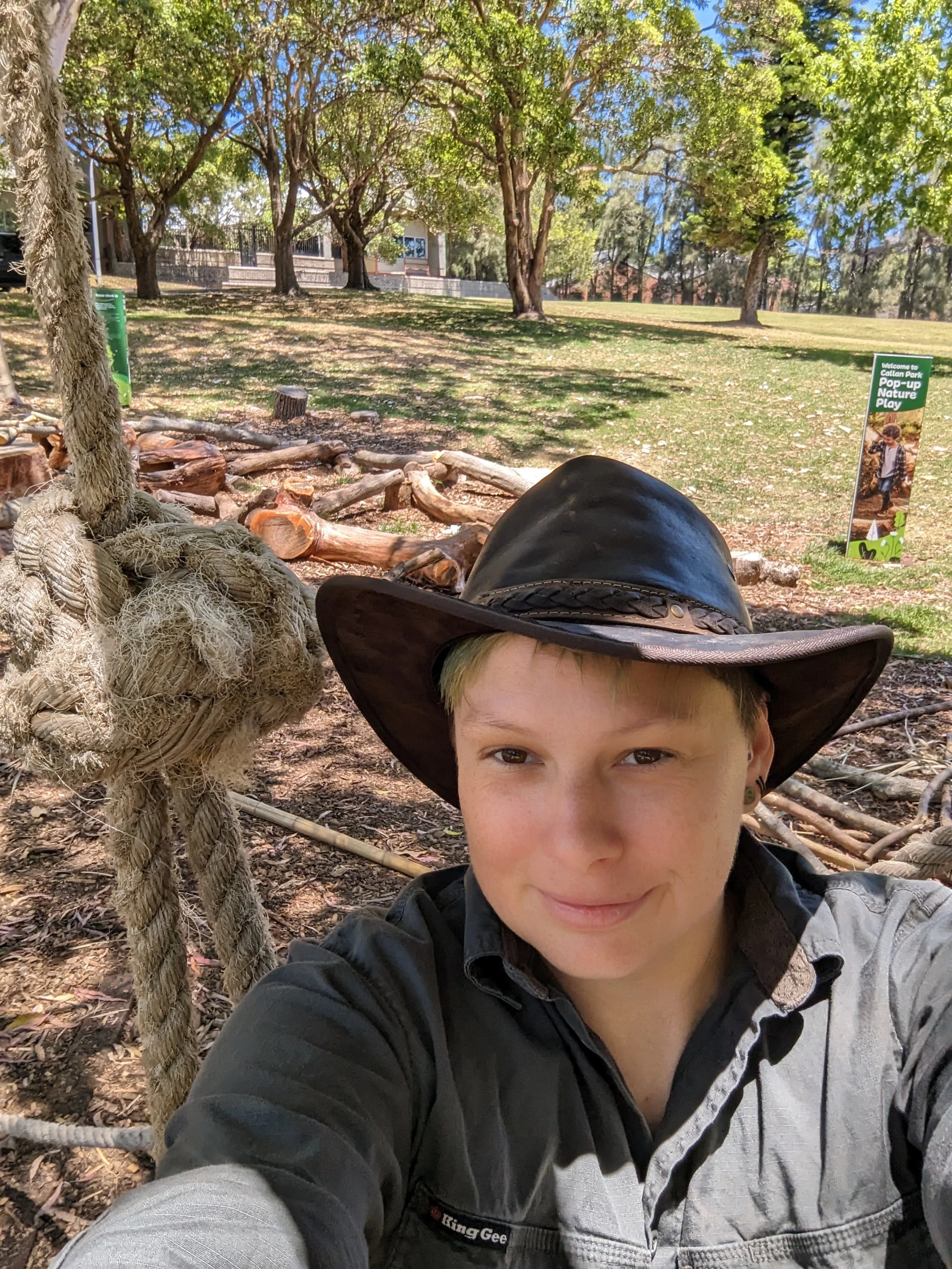 A person taking a selfie outdoors in a park, wearing a wide-brimmed hat and a jacket, with trees, logs, and a sign in the background.
