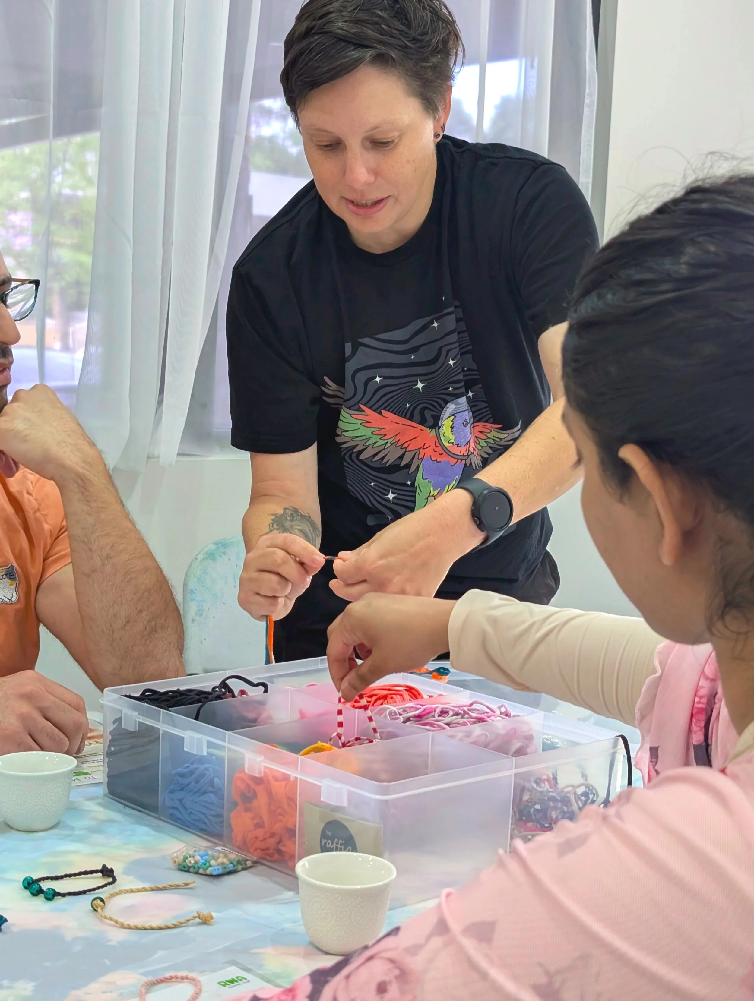 A person in a black graphic t-shirt demonstrates a cord-weaving technique to two participants at a brightly lit workshop table.