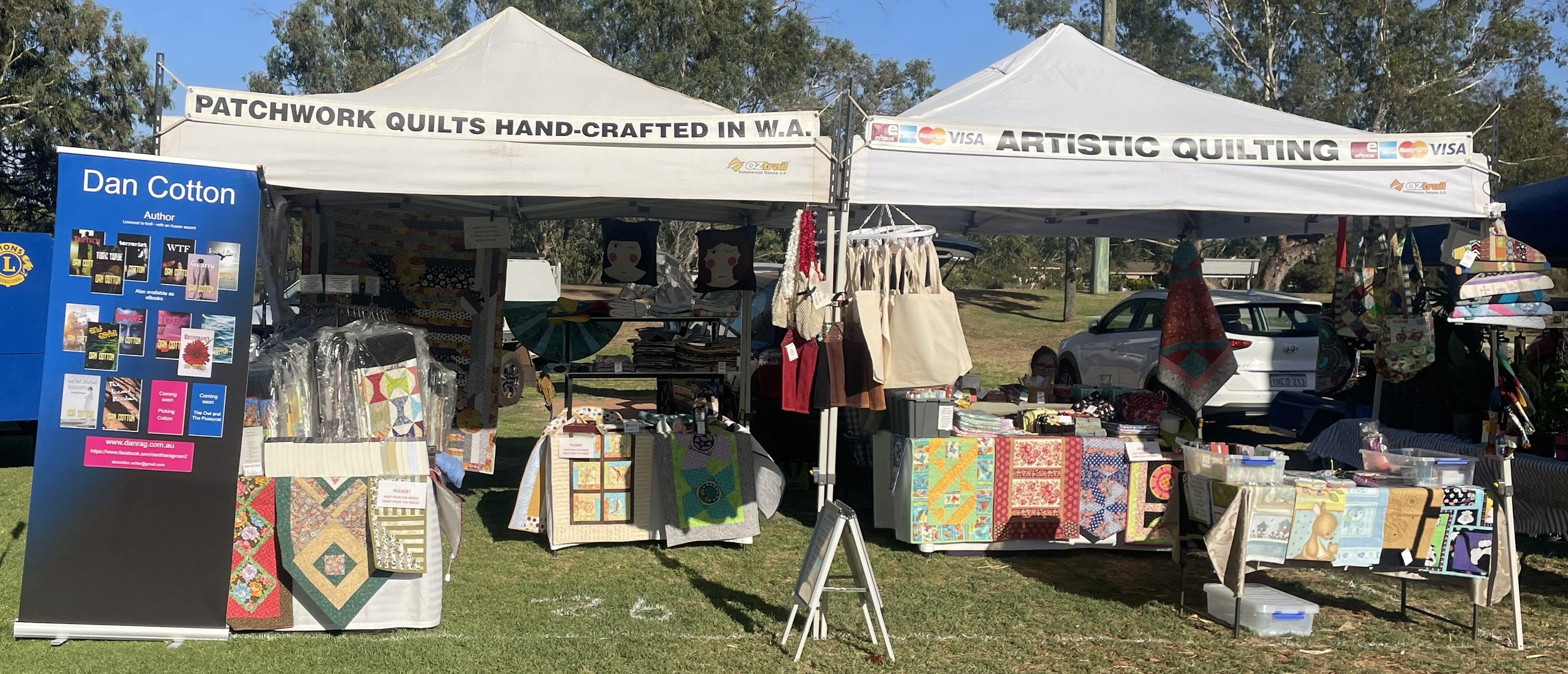 Outdoor craft market stall with quilted items, bags, and fabric crafts under a white tent with signs about patchwork quilts and artistic quilting, cars parked behind, and a person visible sitting at the stall.