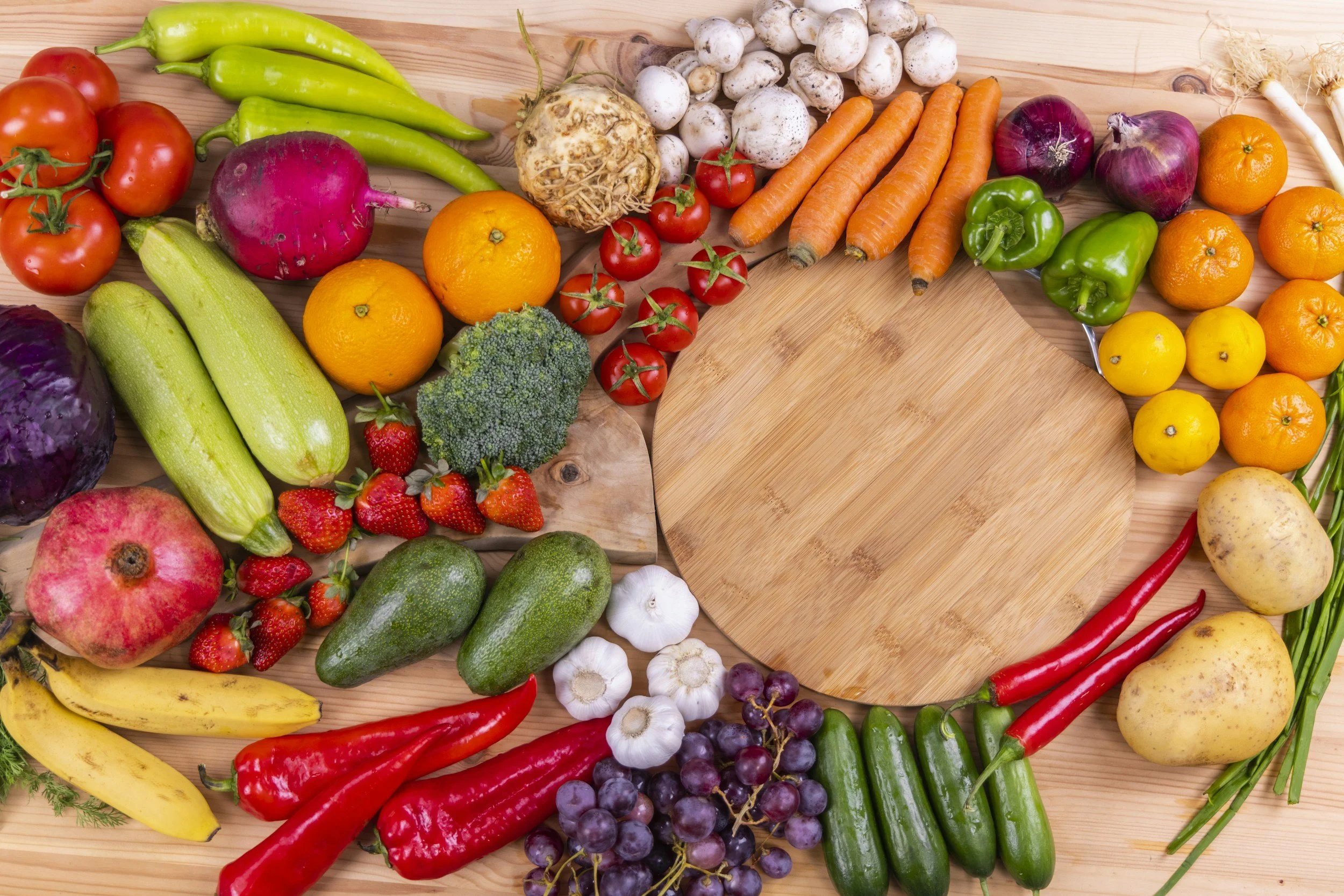 Assorted fresh vegetables and fruits arranged on a wooden surface with a round wooden cutting board in the center, including tomatoes, green peppers, carrots, mushrooms, potatoes, cucumbers, broccoli, strawberries, bananas, grapes, red onions, garlic, and an orange.
