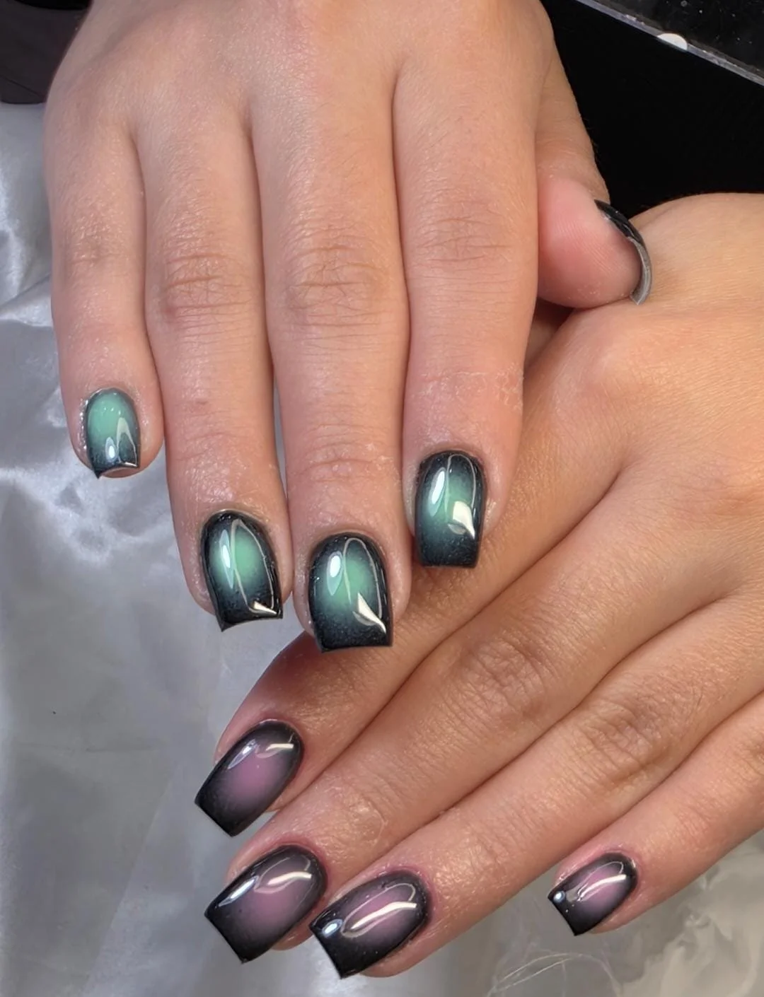 Close-up of manicured fingernails with an ombre gradient design, transitioning from black to green on some fingers and black to pink on others.