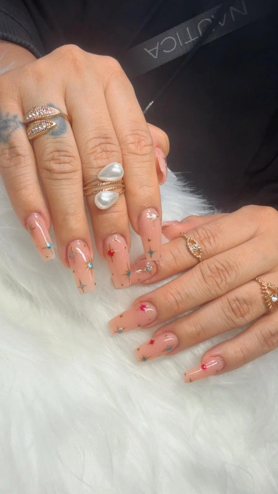 Hands with colorful star and dot nail art, multiple rings, and tattoos, resting on a white furry surface.