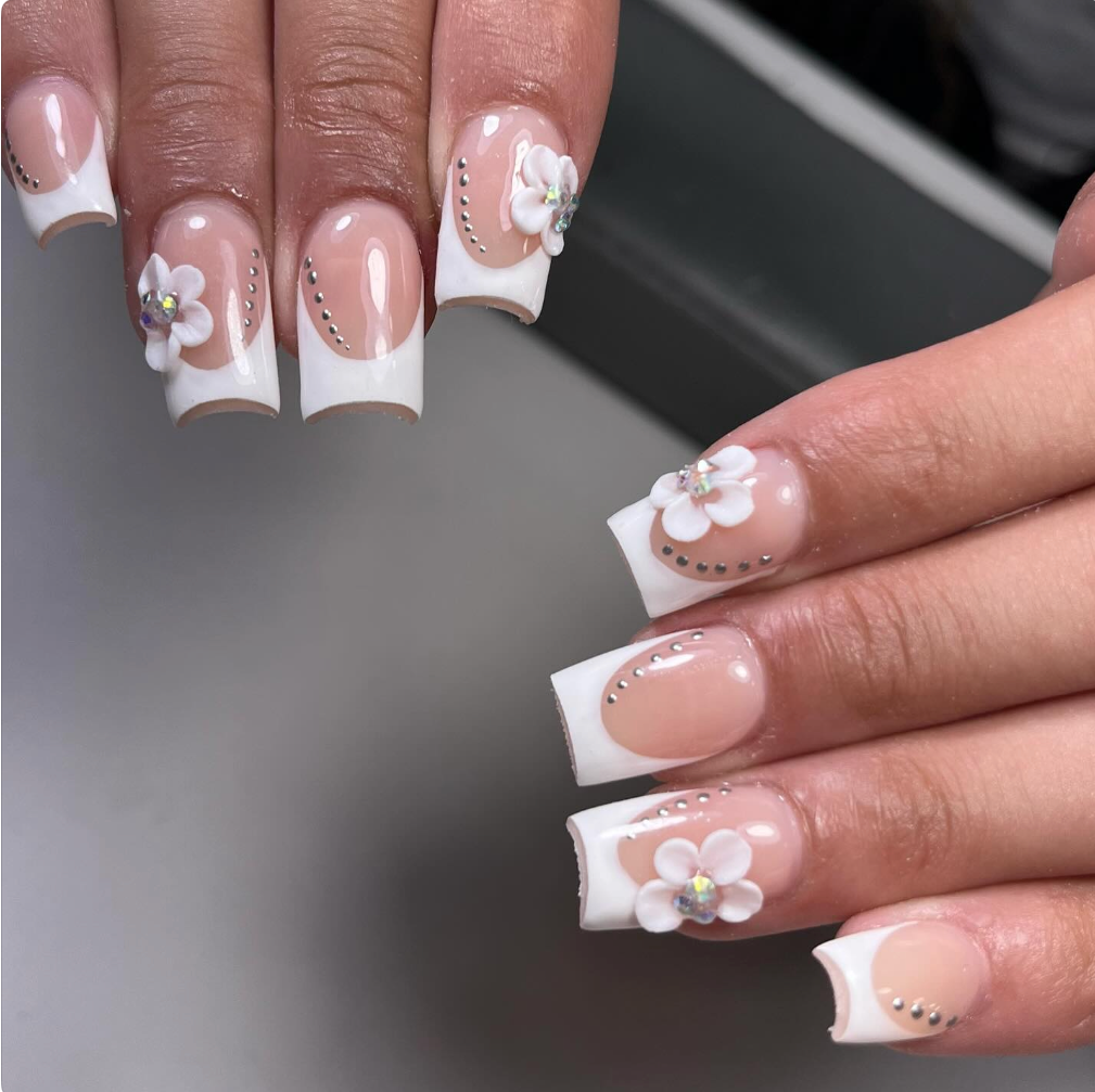 Close-up of manicured nails with white French tips, decorated with 3D white flower embellishments and rhinestones, on a person's hands.