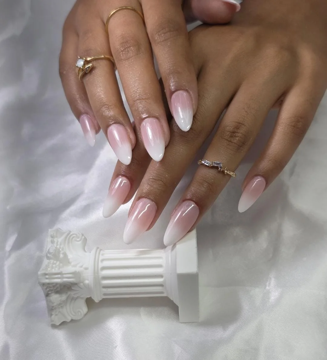 Close-up of a woman's hands with well-manicured, long, white-ombre nails, wearing gold rings, resting on a satin surface near a white decorative column.