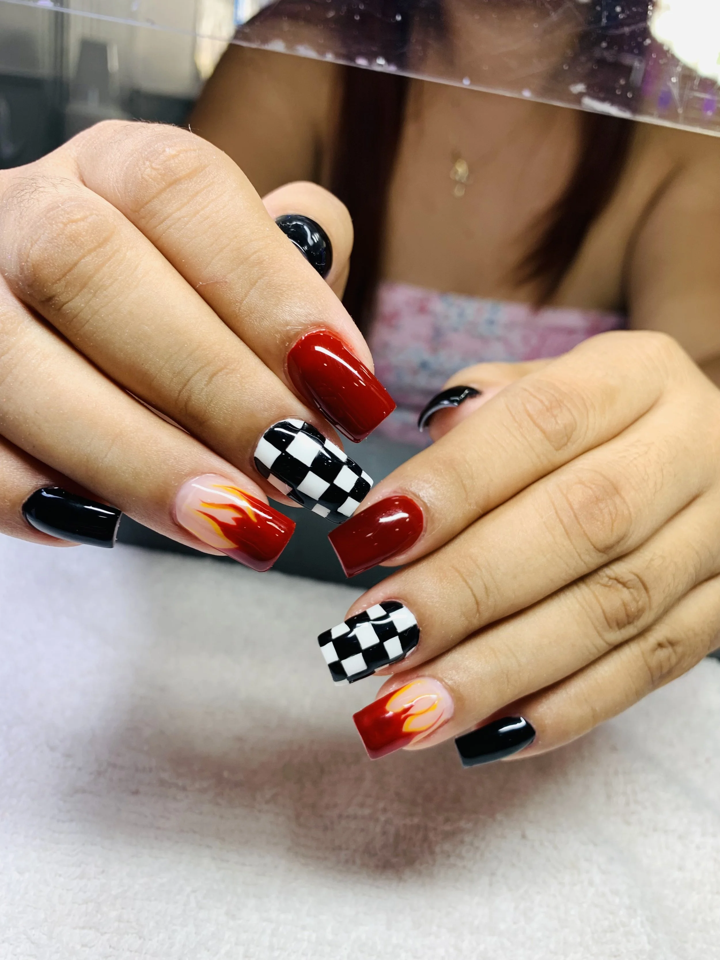 Close-up of a person's hand showing brightly decorated fingernails featuring black, red, checkered patterns, and flames, with part of their shirt and a glass barrier in the background.