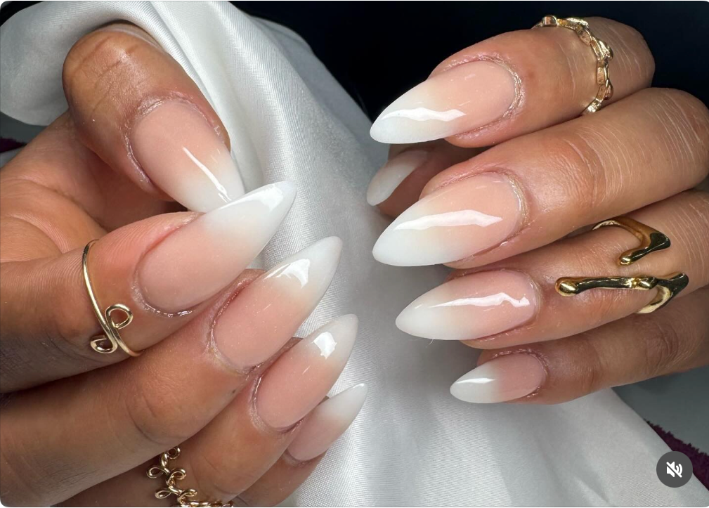 Close-up of hands with long, almond-shaped, white-to-clear ombre acrylic nails, adorned with gold jewelry.