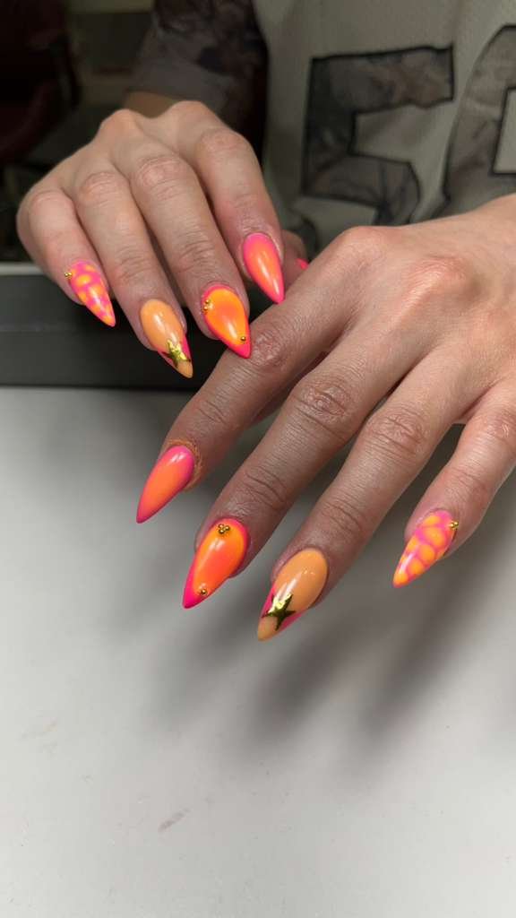 Close-up of manicured hands with vibrant orange and pink gradient nails featuring floral and geometric designs, small gold accents, and embellishments.