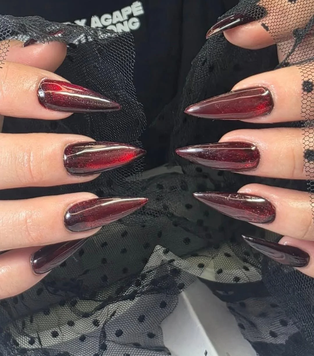 Close-up of long, pointed, metallic red and black nails with a shiny finish, held against black polka dot tulle fabric.