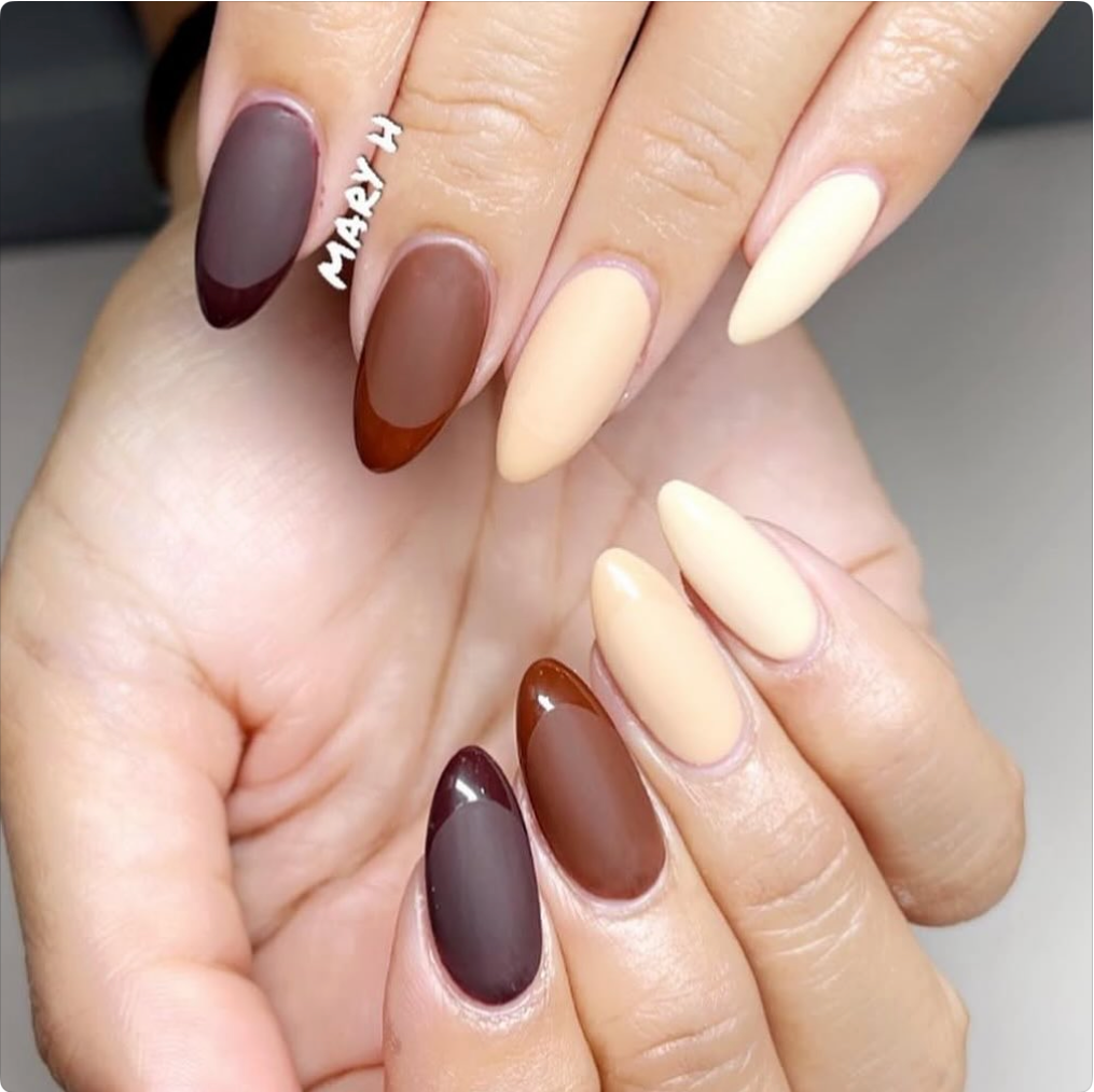 Close-up of hands with almond-shaped nails painted in matte cream, brown, and dark burgundy colors, with some nails featuring a two-tone design.
