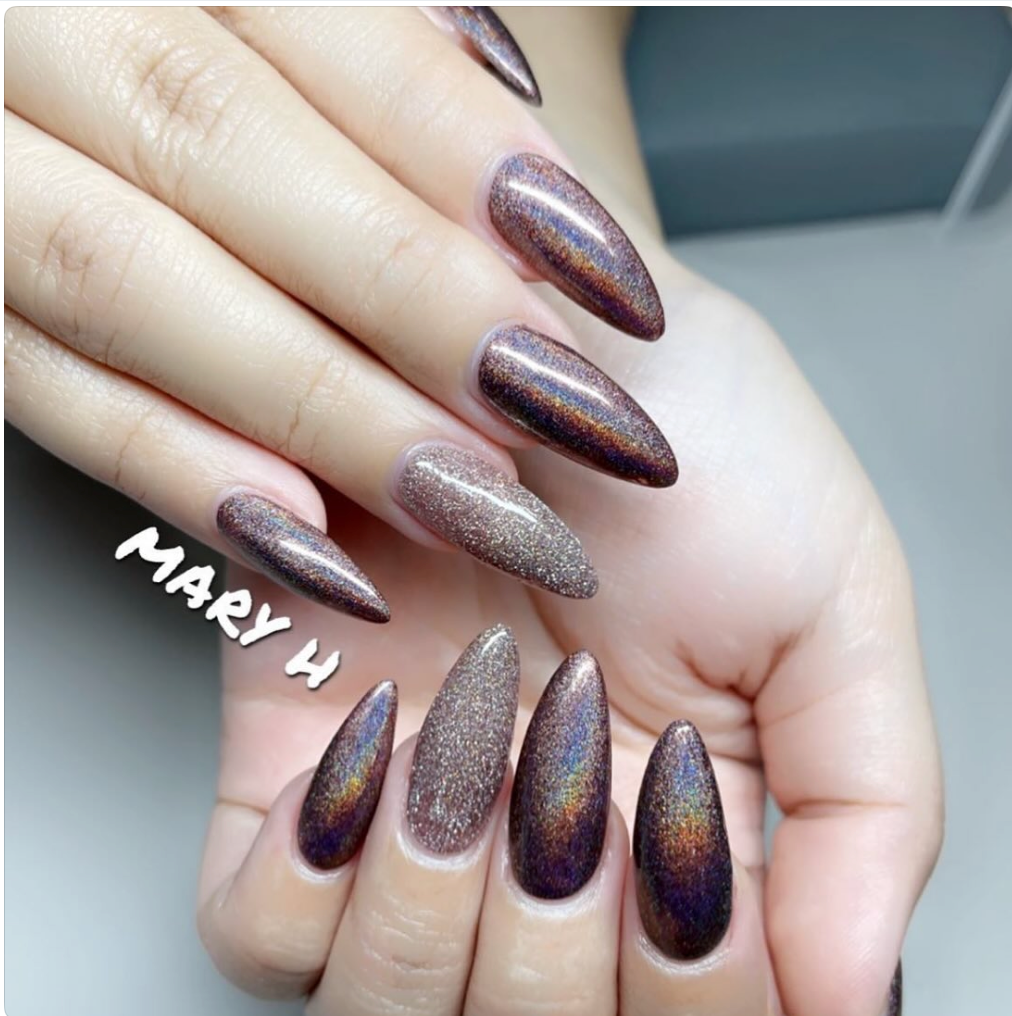 Close-up of a hand with long, almond-shaped, multicolored metallic and glittery nail polish on each fingernail. The nails feature a shiny finish with a rainbow-like sheen and the text 'MARY H' written in white on the image.