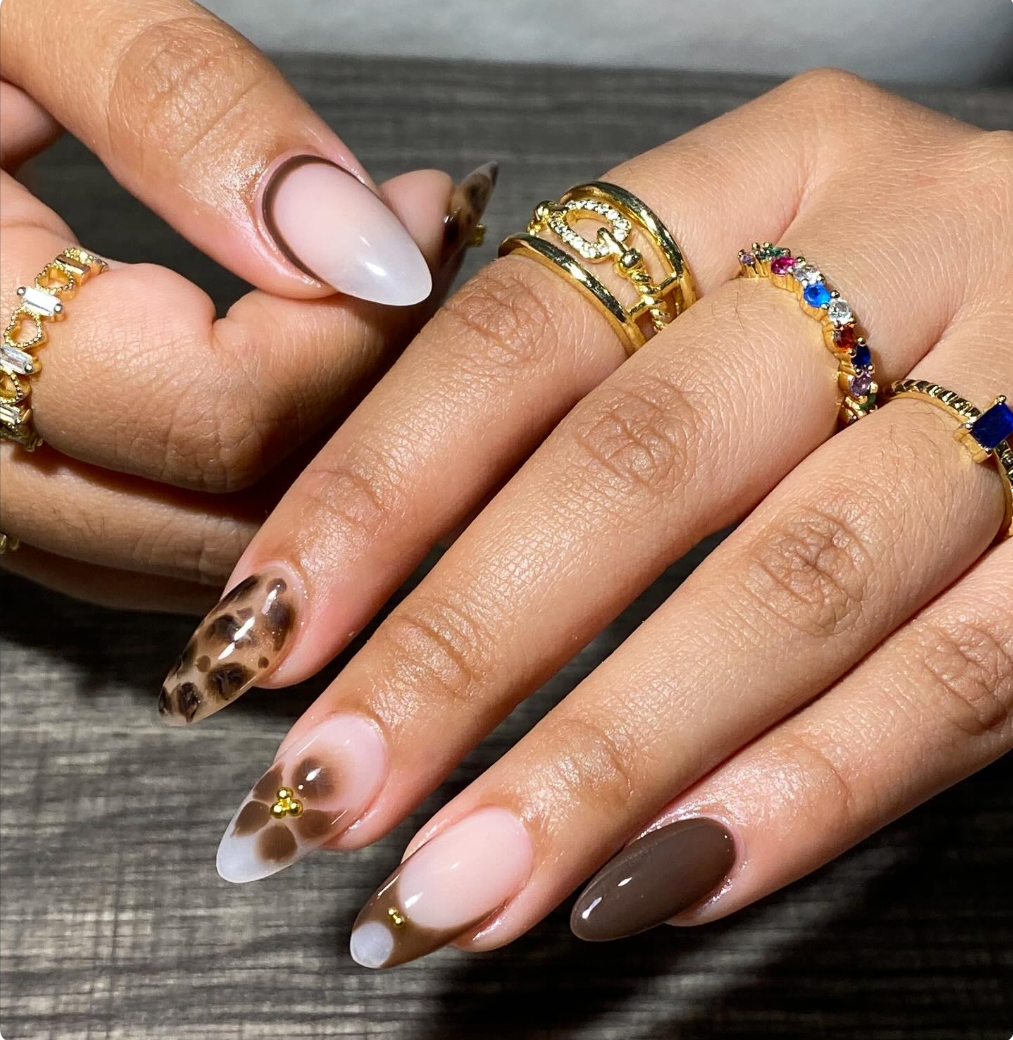 A hand with decorated nails, featuring a marble and floral design with small gold embellishments, and multiple gold rings with various gemstones.