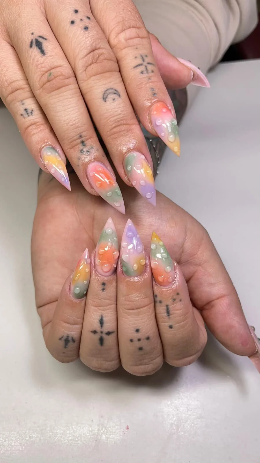 Close-up of hands with colorful, rainbow-themed nail art featuring pastel shades and bubble designs, and various small black tattoos on fingers.