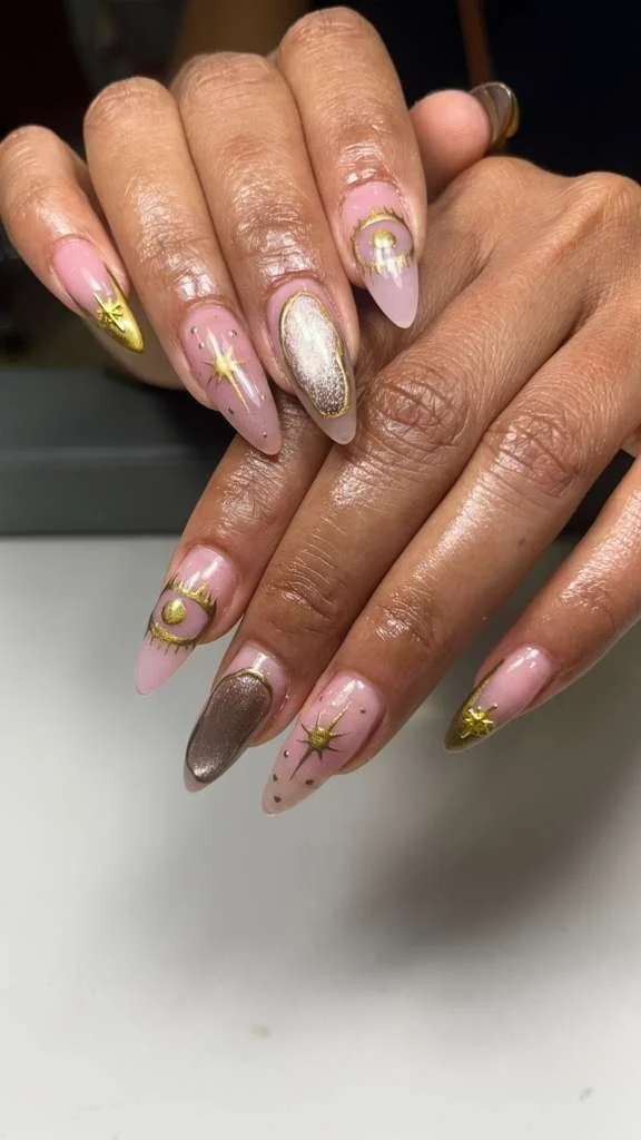 Close-up of a person's manicured nails with pink and gold nail polish, adorned with gold celestial and star designs.