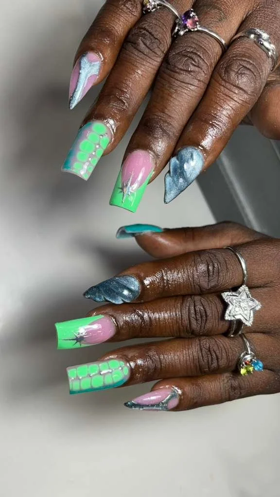 Close-up of hands with colorful, decorated acrylic nails featuring neon green, pink, blue, and white designs, along with multiple rings.