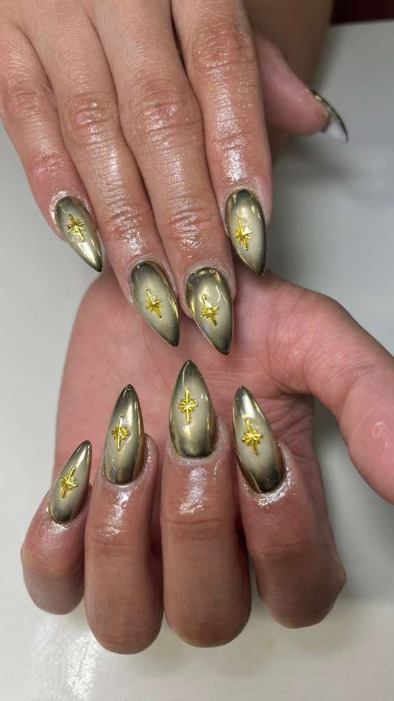 Close-up of a person's manicured nails with metallic gold and black ombré design, small gold star and cross ornaments on each nail.