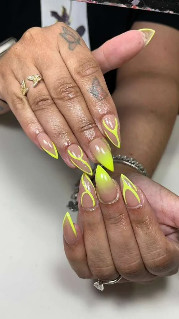 A close-up of a person's hands with long, neon yellow and clear acrylic nails with abstract designs. The person has tattoos on their fingers and multiple rings, including a silver band and a ring with a gemstone.