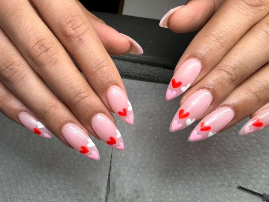 Close-up of hands with pink almond-shaped nails decorated with red, white, and pink heart designs.
