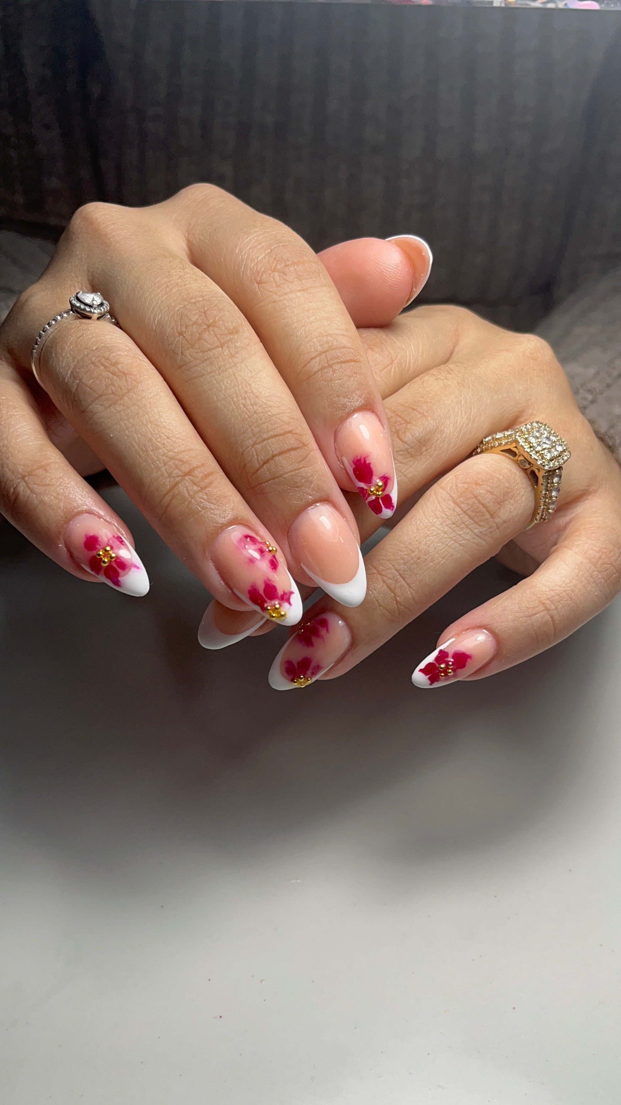 Close-up of hands with manicured nails featuring pink and white floral nail art and gold embellishments. The person is wearing two rings—one silver on the index finger and one gold with diamonds on the ring finger.