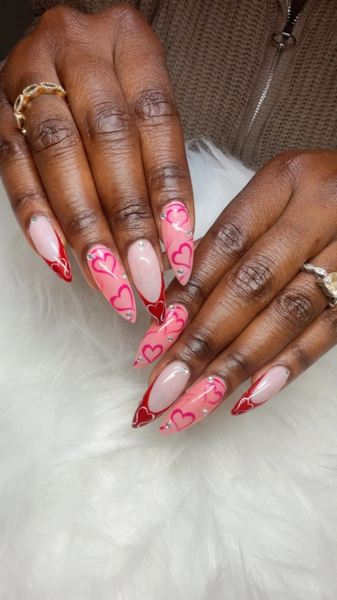 Close-up of a person's hands with Valentine's Day-themed nail art featuring pink hearts, red accents, and rhinestones.