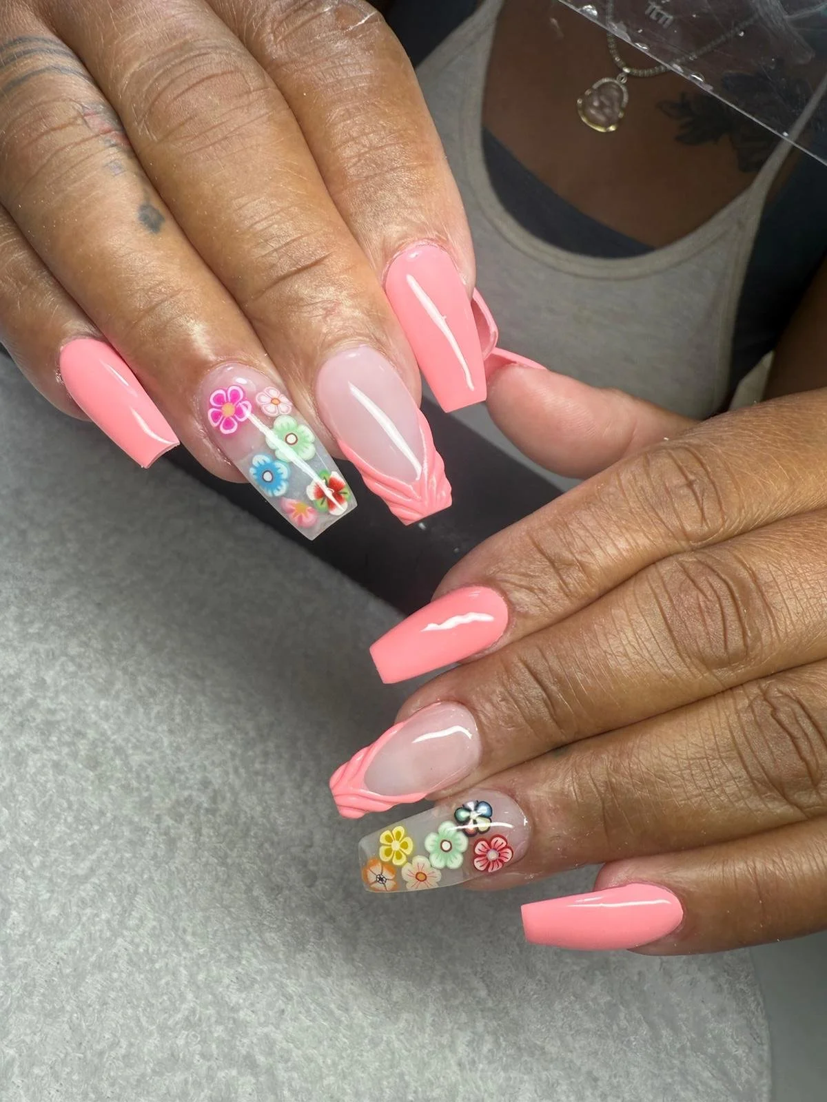 Close-up of hands with pink and floral decorated acrylic nails, showing some nails with solid pink polish, some with a clear base and colorful flower designs, and one with pink nail art.
