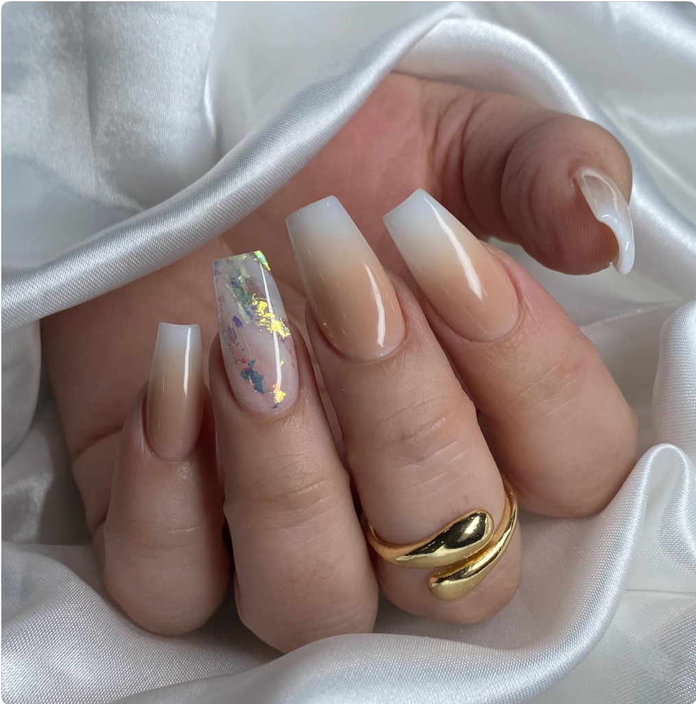 Close-up of a hand with manicured nails, featuring a nude and white gradient design on most nails and an accent nail with iridescent holographic foil. The hand is adorned with a gold ring and rests on white satin fabric.