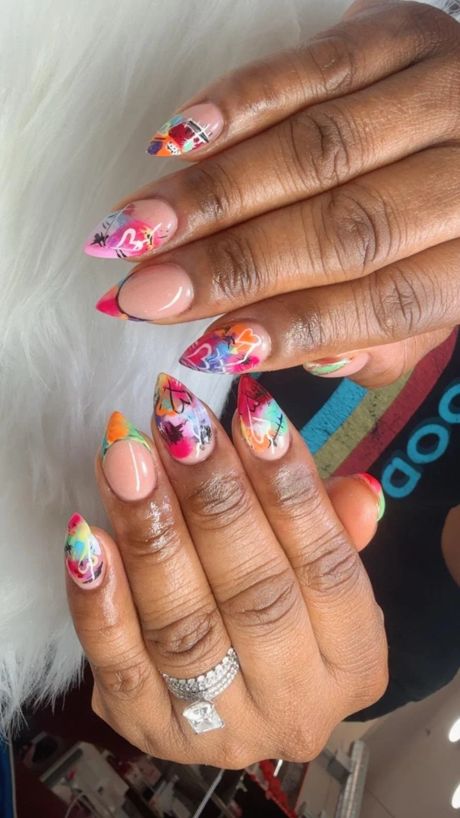 Close-up of a person's hand with multicolored, artistically painted nails featuring abstract designs and hearts. The individual is wearing a diamond ring on their finger.