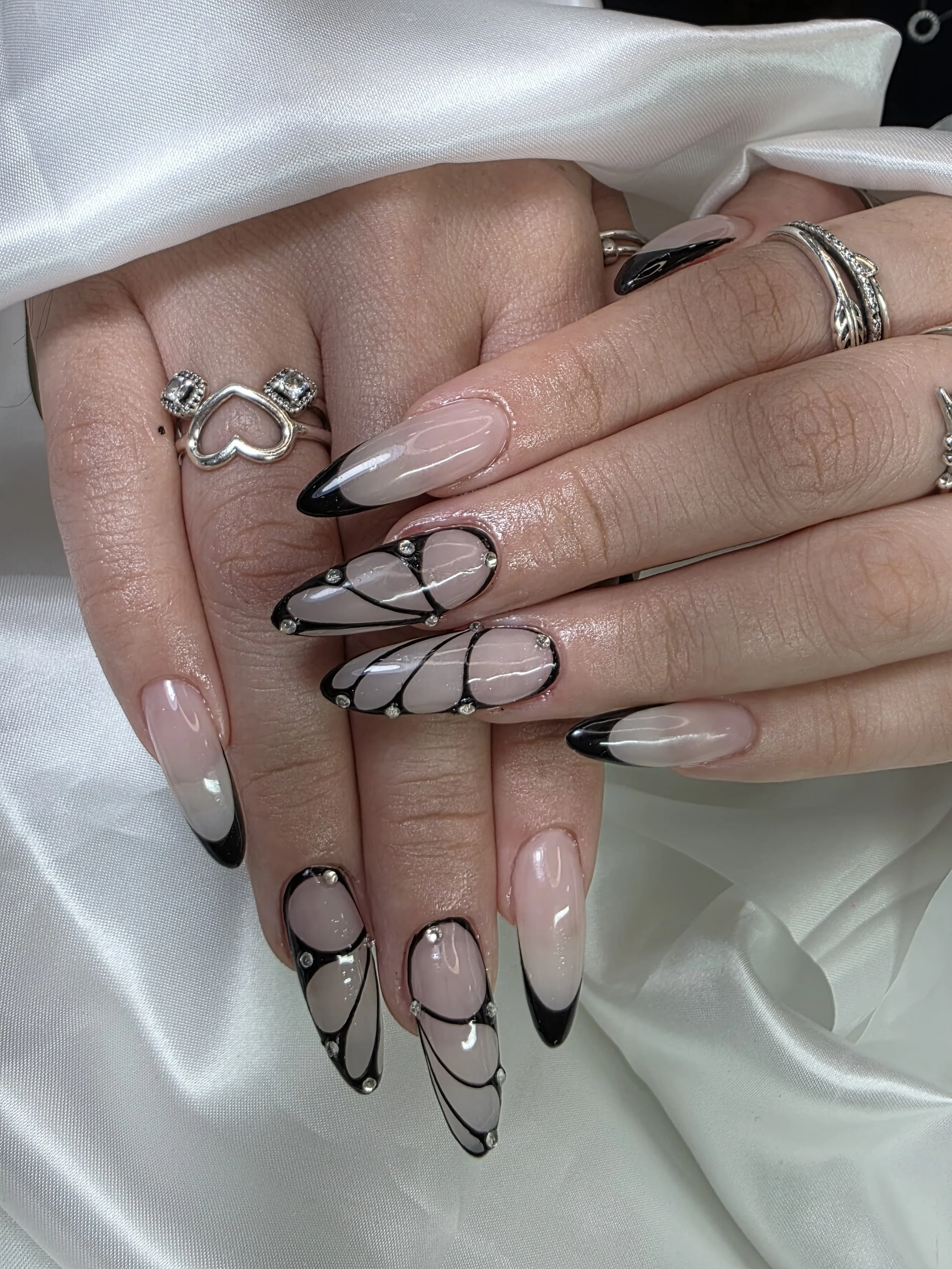 Close-up of hands with manicured nails, featuring black and beige nail designs and rings, resting on white satin fabric.