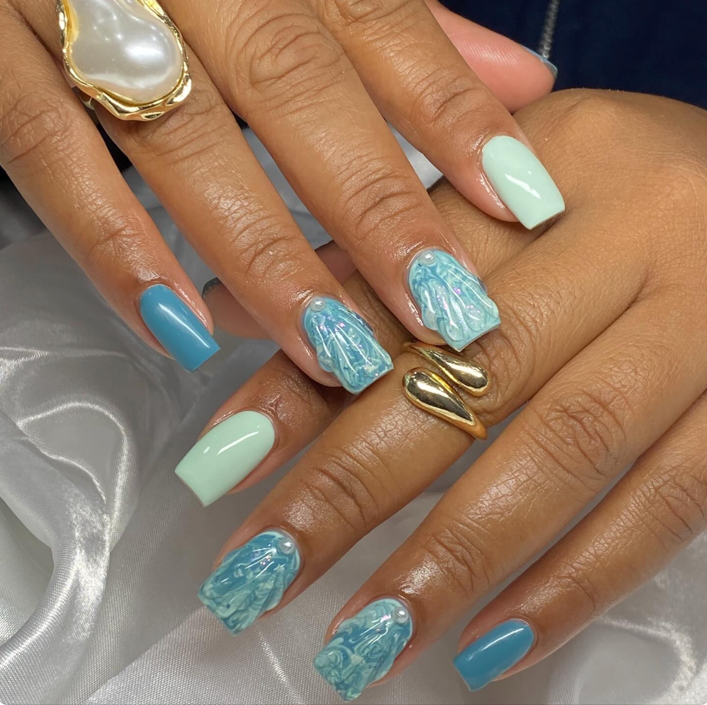 Close-up of a person's hands with pastel-colored nail polish, including blue, mint green, and patterned designs with pearl accents, showing a gold ring with a large pearl.