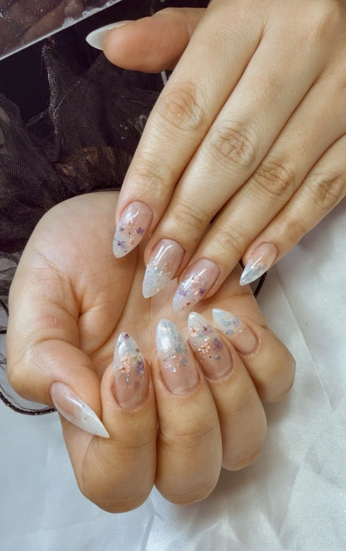 Close-up of hands with decorated acrylic nails featuring a clear base with pastel and glitter accents, positioned on a white satin surface.