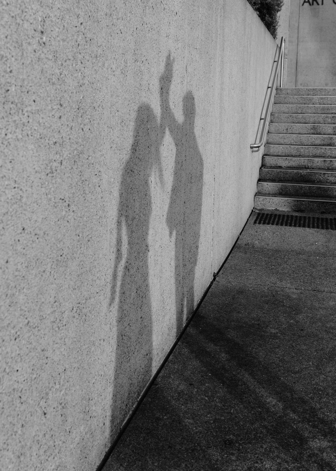 Shadow of two people leaning on a wall, one raising an arm, next to a staircase outside.