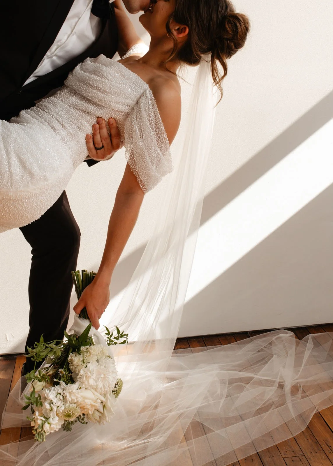 A bride holding a bouquet of white flowers, leaning backward with her eyes closed, while a groom holds her and kisses her forehead. The bride is wearing a beaded wedding gown with a tulle train, and the groom is in a black tuxedo with a white shirt. They are in a softly lit room with a plain white wall in the background.