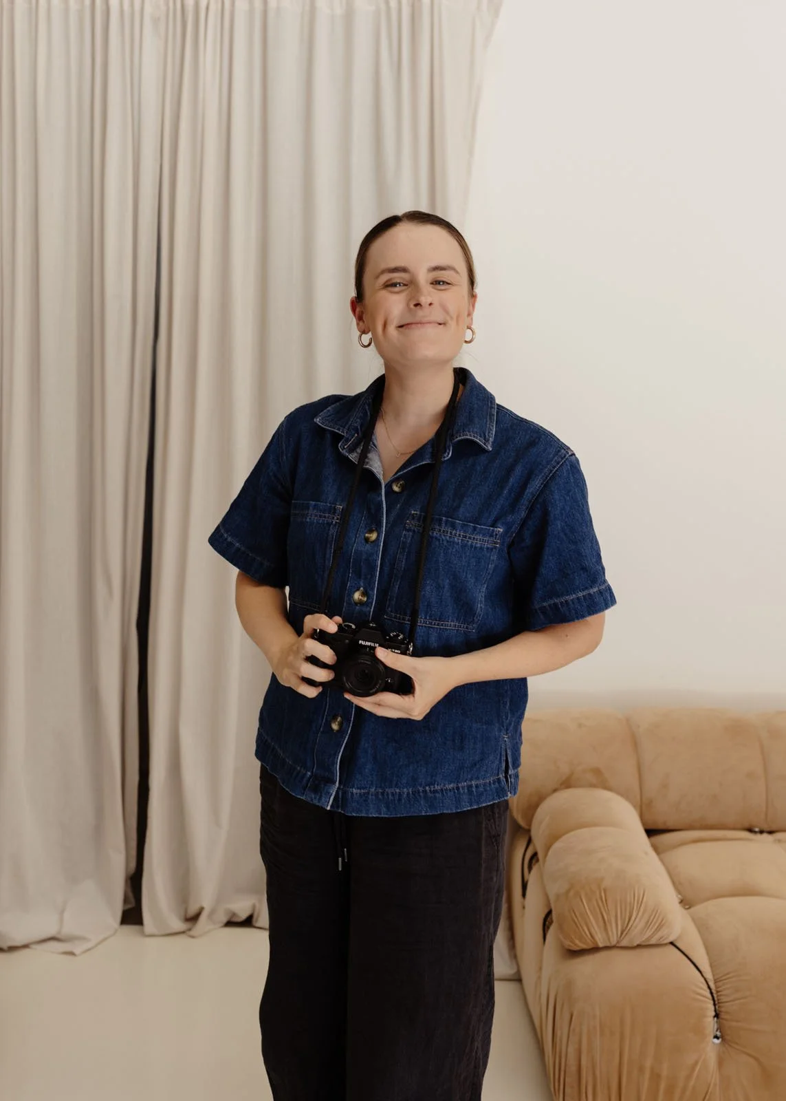 Woman with short brown hair smiling, wearing a denim jacket and black pants, holding a camera, standing in a room with beige Curtains and a beige couch.