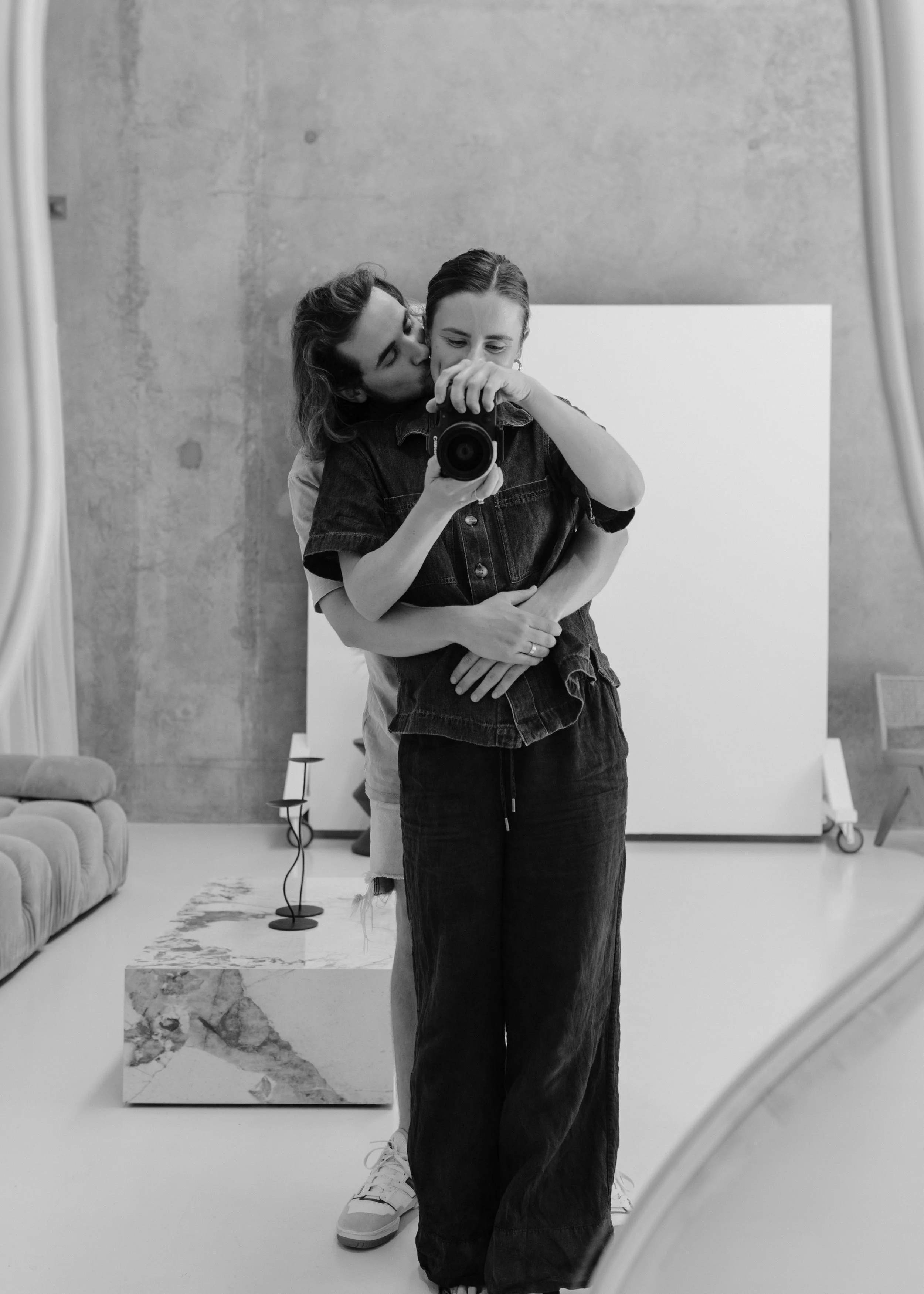 A couple taking a mirror selfie in a studio. The man is kissing the woman on the cheek from behind, and they are both smiling lightly. The woman is holding the camera, and they are dressed casually.